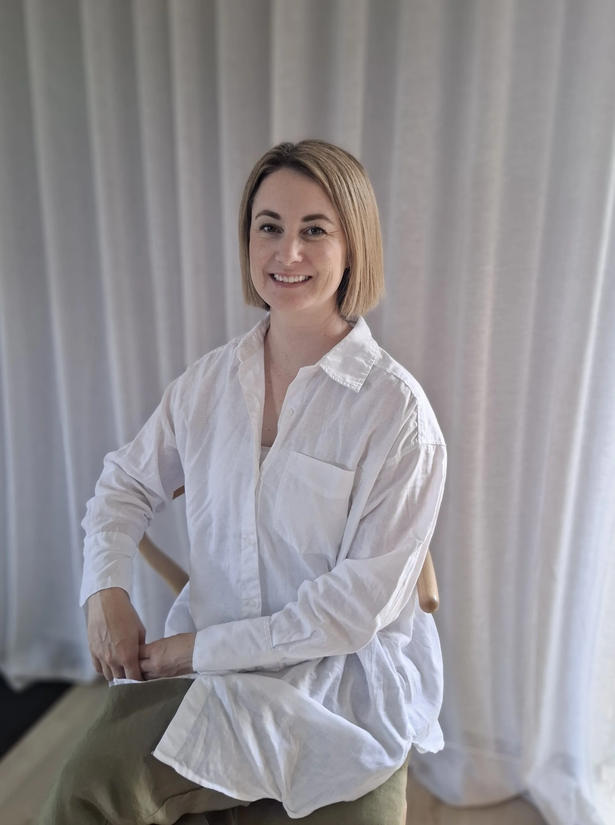 A woman with a short blonde bob haircut sitting on a chair in front of light-colored curtains, smiling at the camera, wearing a loose white button-down shirt and beige pants.