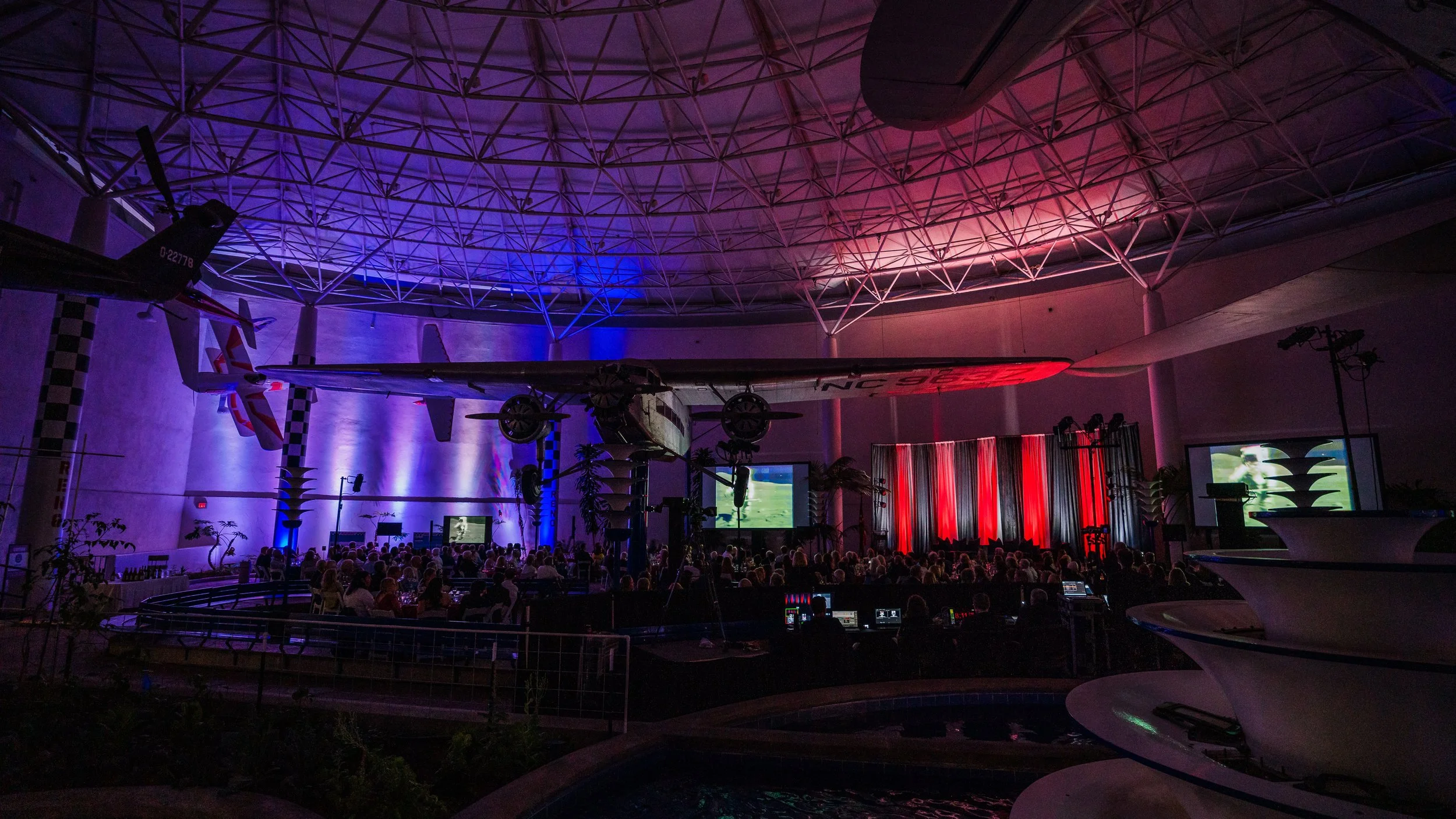 An indoor event space with a large audience, a stage with a red curtain, two screens displaying content, purple and red lighting, and a small airplane suspended from the ceiling.