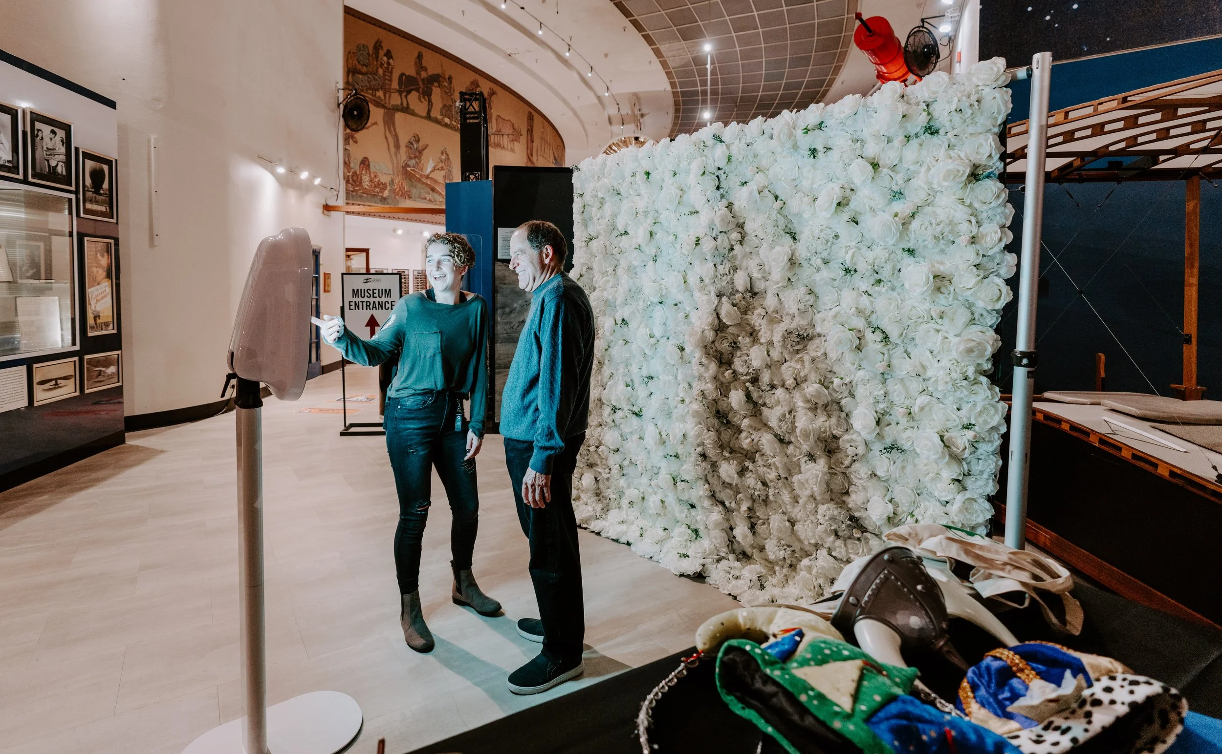Two people standing near a large wall of white roses inside a museum, with a sign indicating the museum entrance behind them. One person is pointing at a device on a stand, and there are various objects and a mask on the table in the foreground.