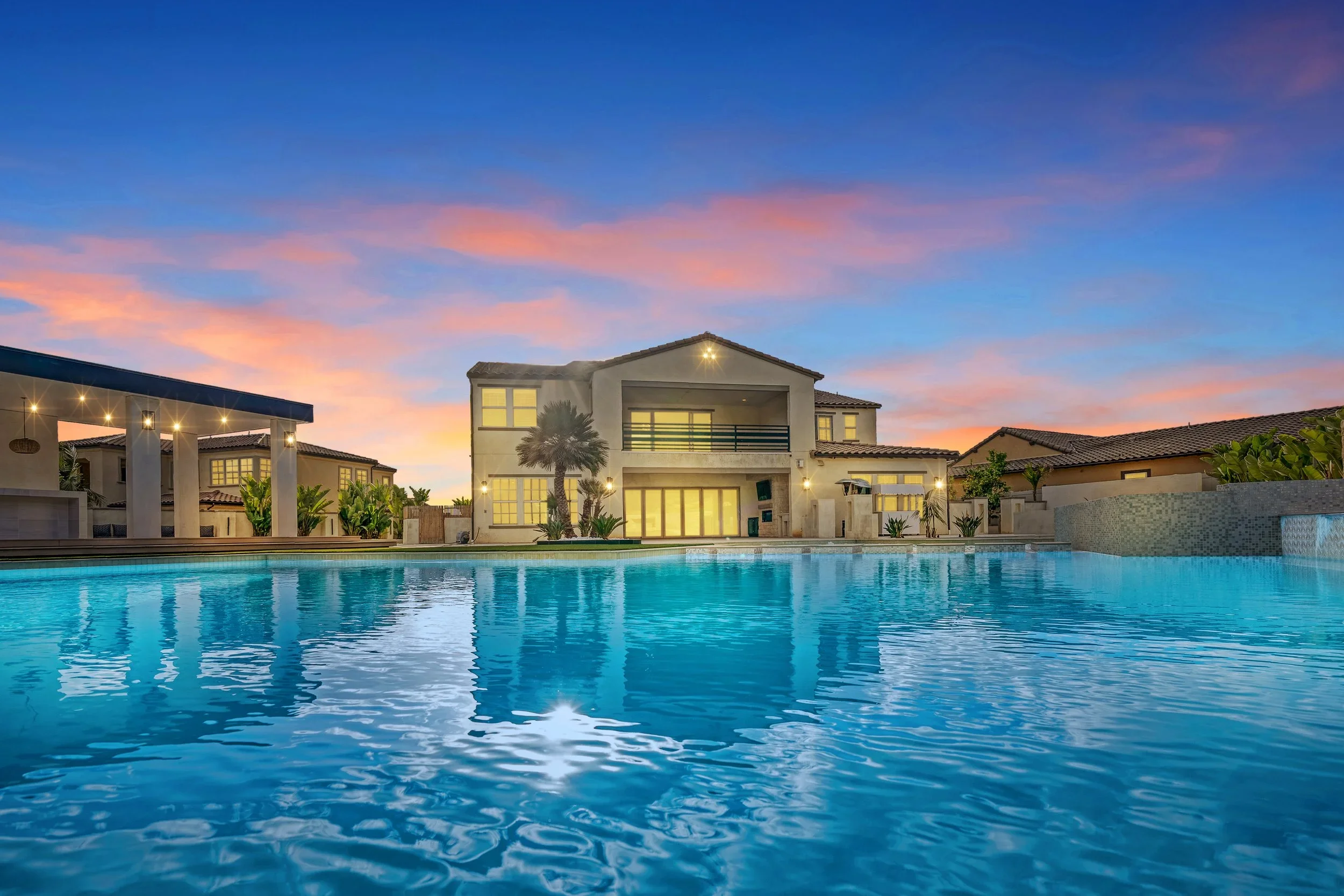 Luxury house with a large swimming pool in front, palm trees, and a vibrant sky at sunset.