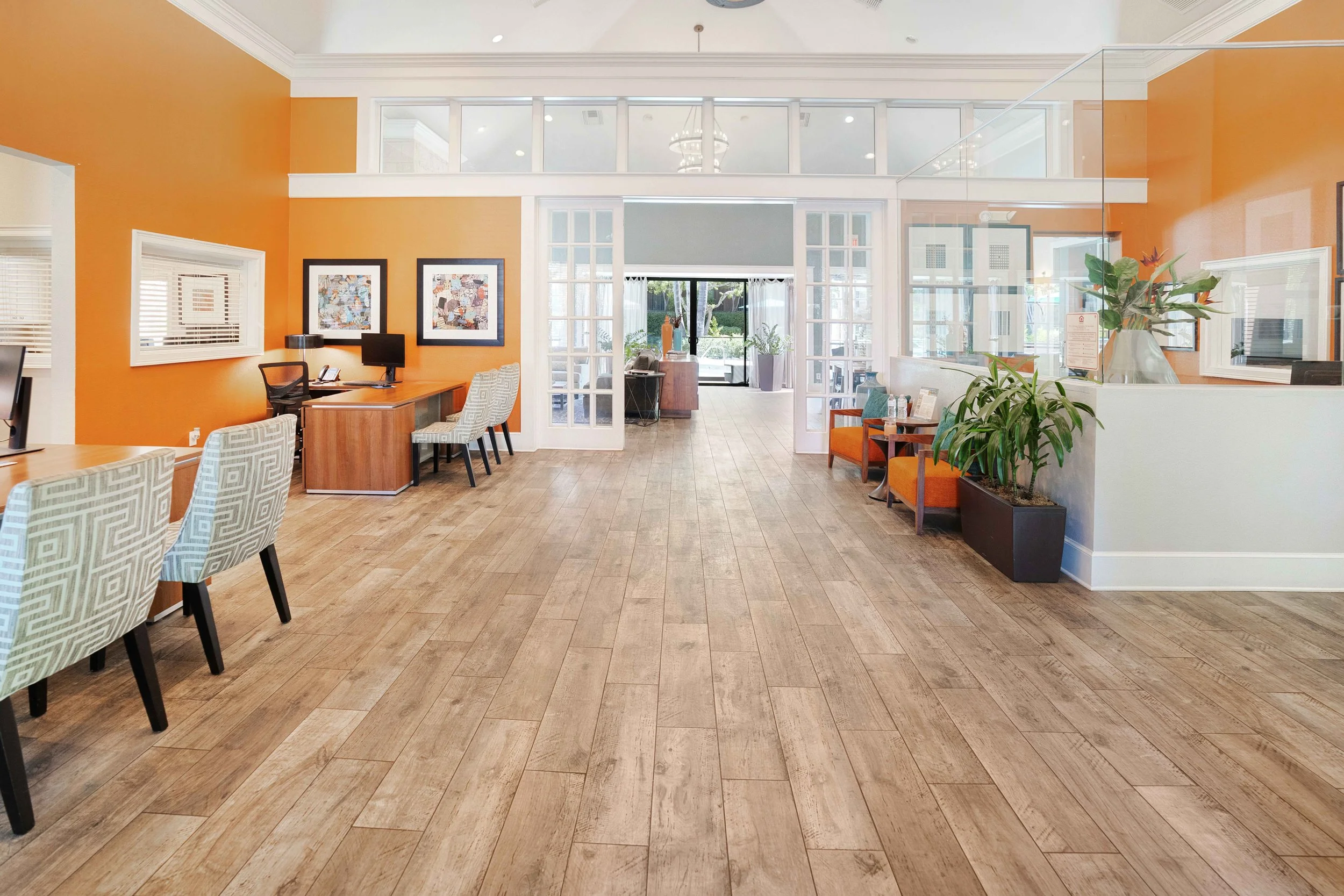 Bright hotel lobby with orange accent wall, wooden floors, seating area with chairs and tables, computer workstations on the left, potted plants, glass doors leading outside, and tasteful artwork on the walls.