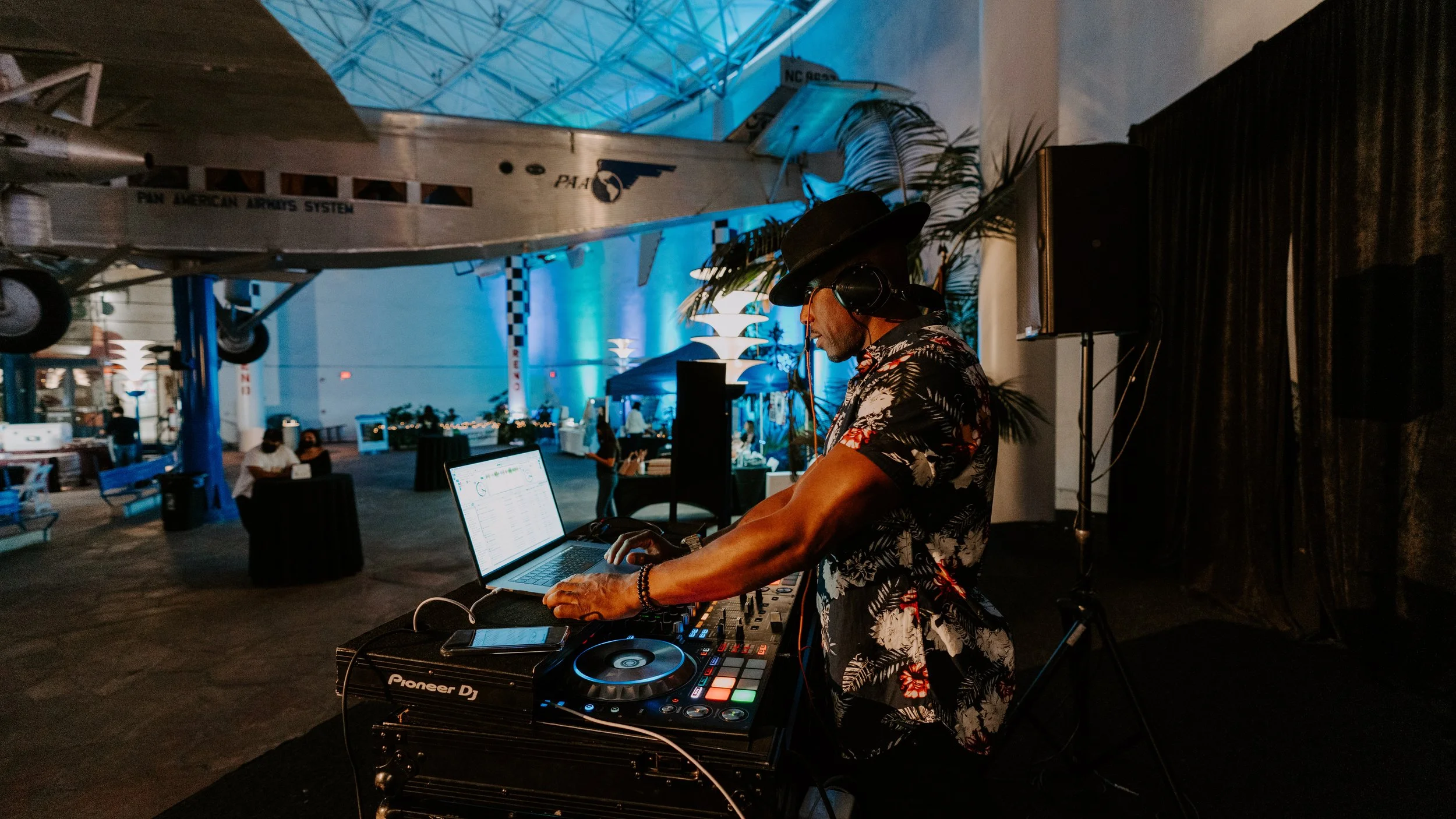 DJ wearing a floral shirt, hat, and headphones, mixing music at an indoor event with a DC-3 airplane overhead, dark lighting, and blue accent lights.