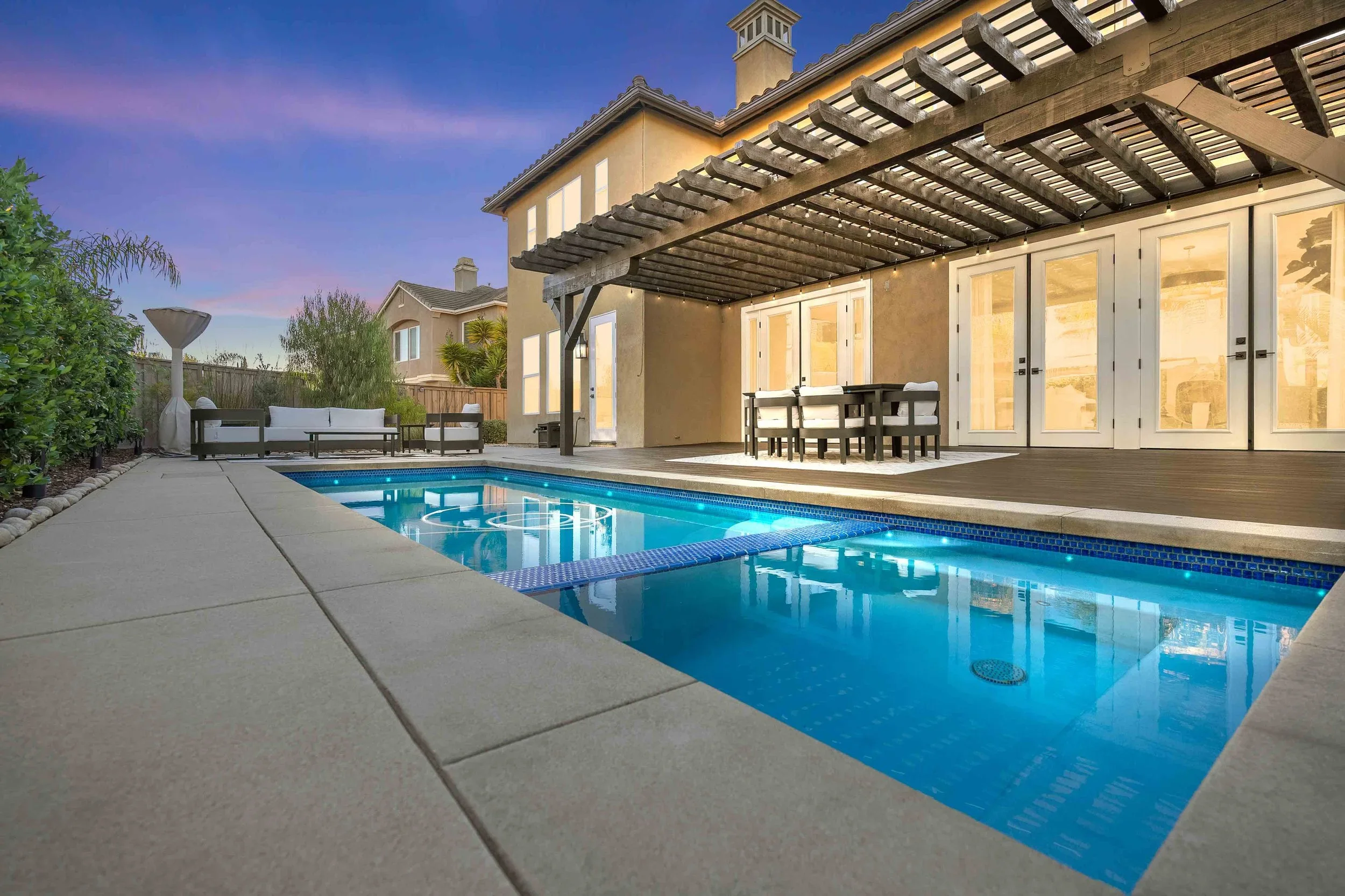 A backyard with a rectangular swimming pool, outdoor seating, a wooden deck, a beige house with large glass doors, a pergola, and landscaped greenery under a sky transitioning from pink to blue.