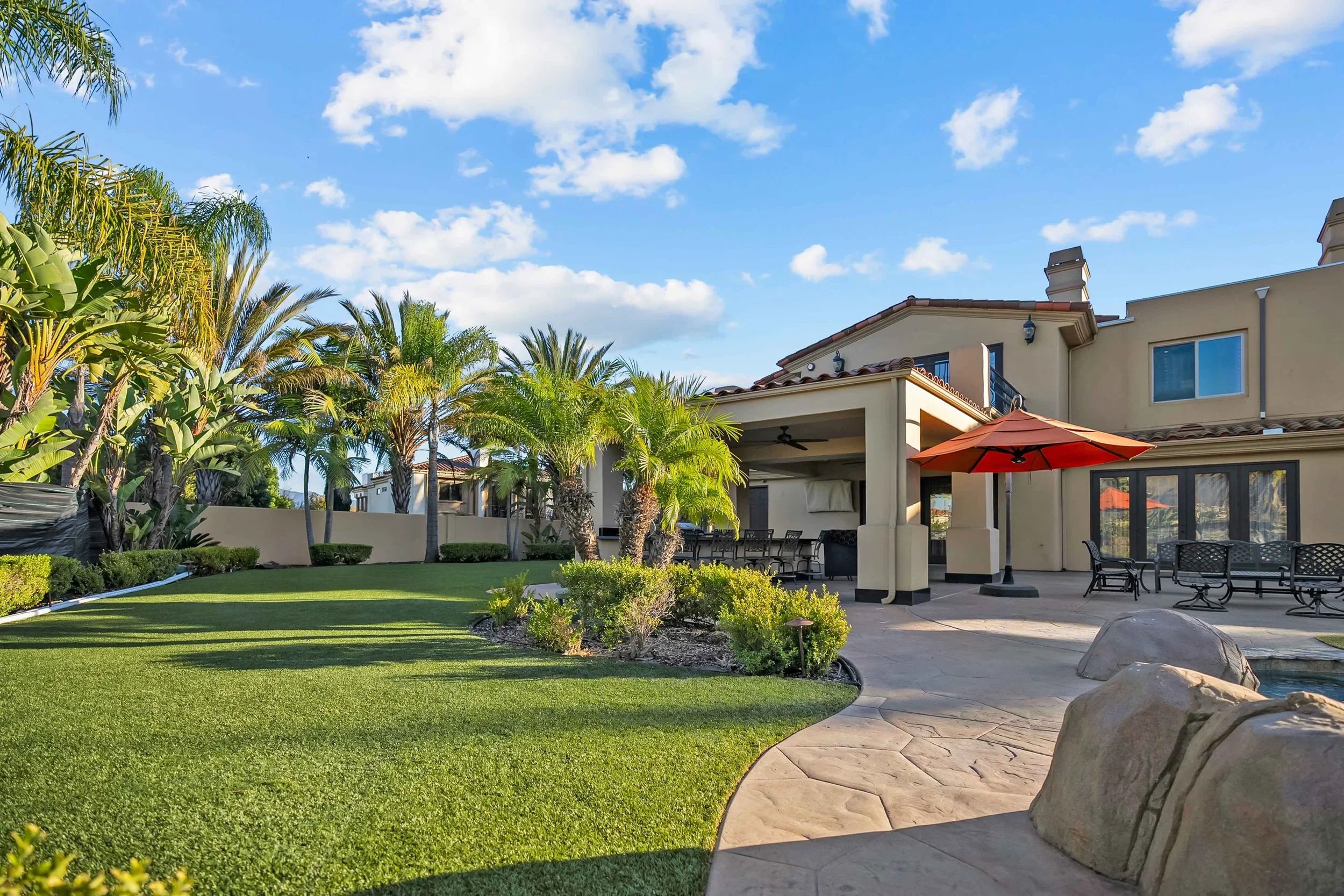 A backyard with lush green grass, palm trees, and an outdoor patio area with tables, chairs, and an umbrella on a sunny day.