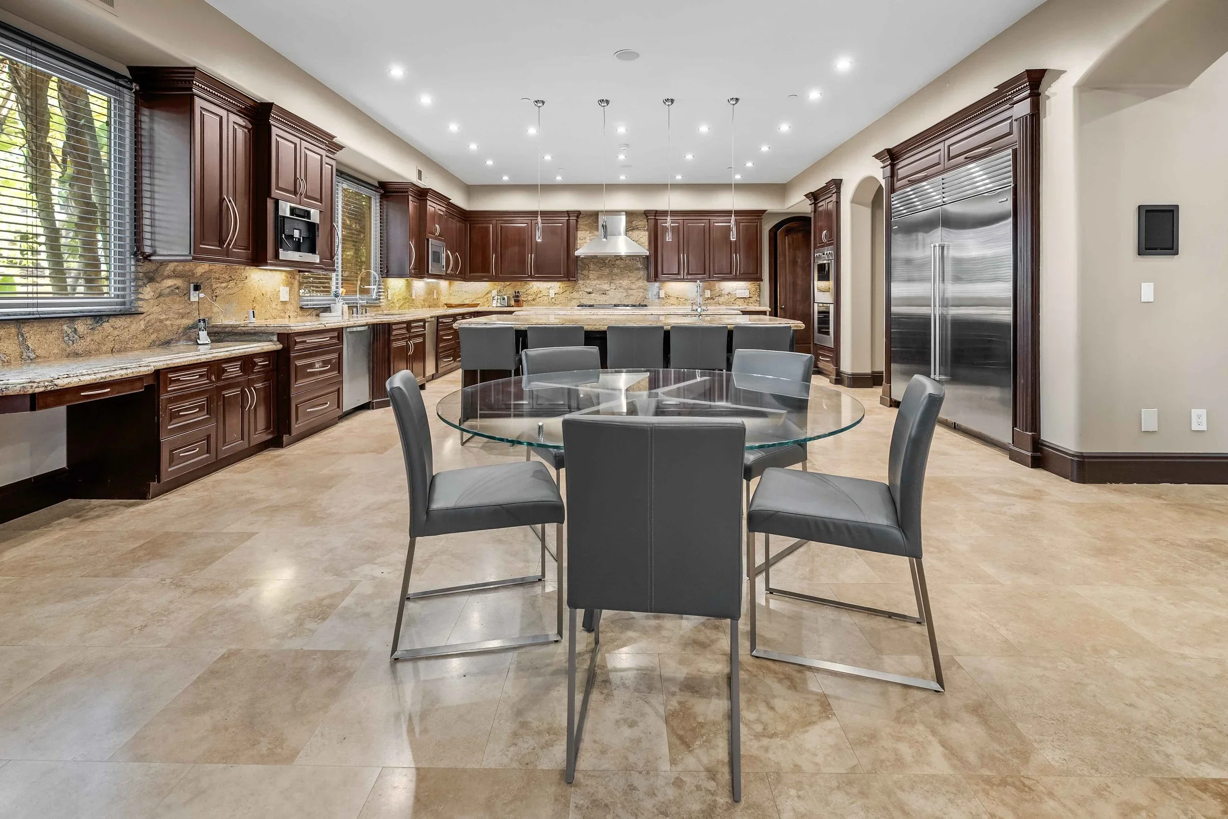 Modern kitchen with wooden cabinets, granite countertops, stainless steel appliances, and a round glass dining table with six gray chairs.