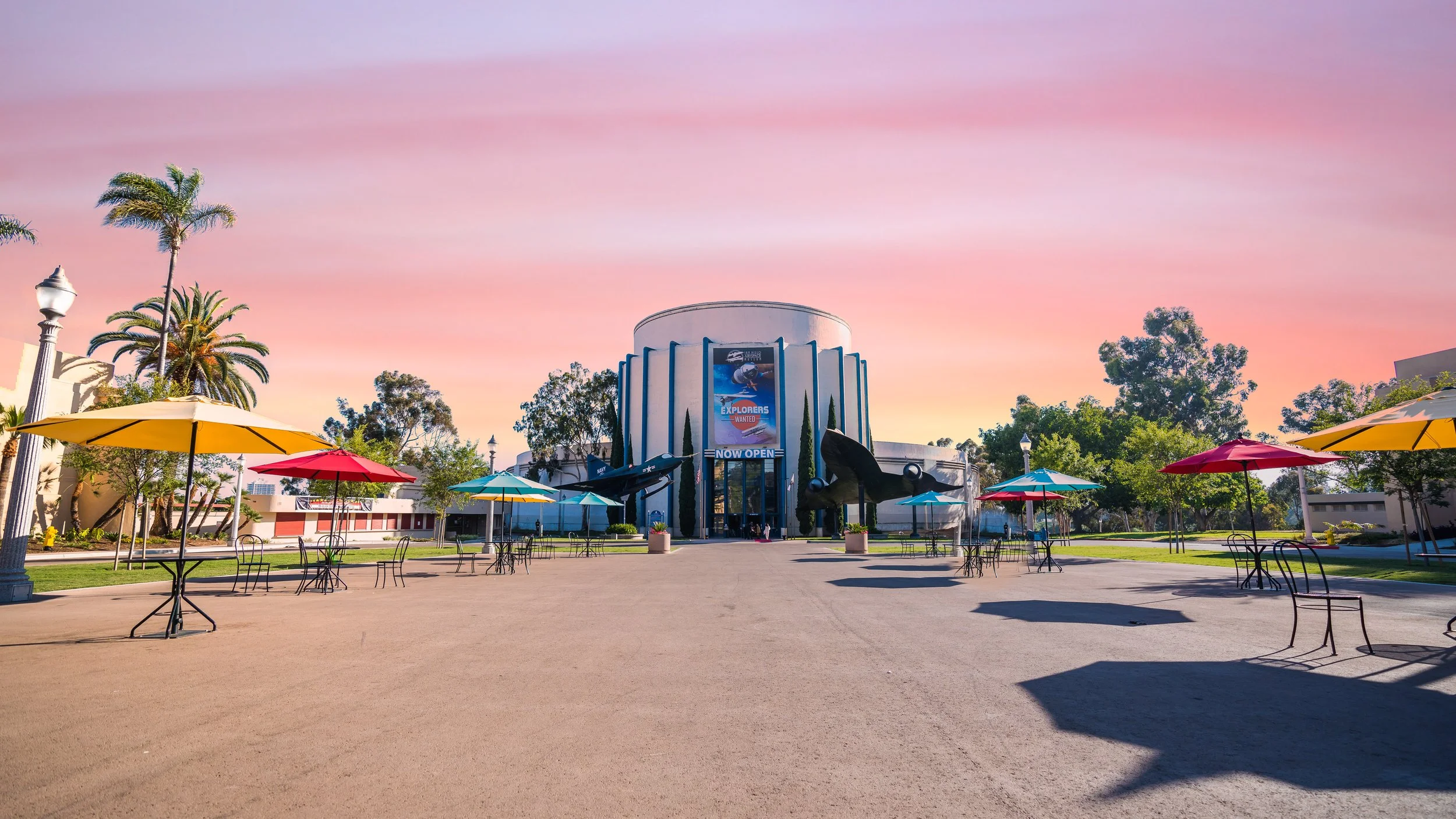 Large outdoor area with colorful umbrellas and tables in front of a museum with a space exploration theme. The museum has a sign that reads "Now Open" and displays a large banner and two jet aircraft on either side of the entrance. There are trees an