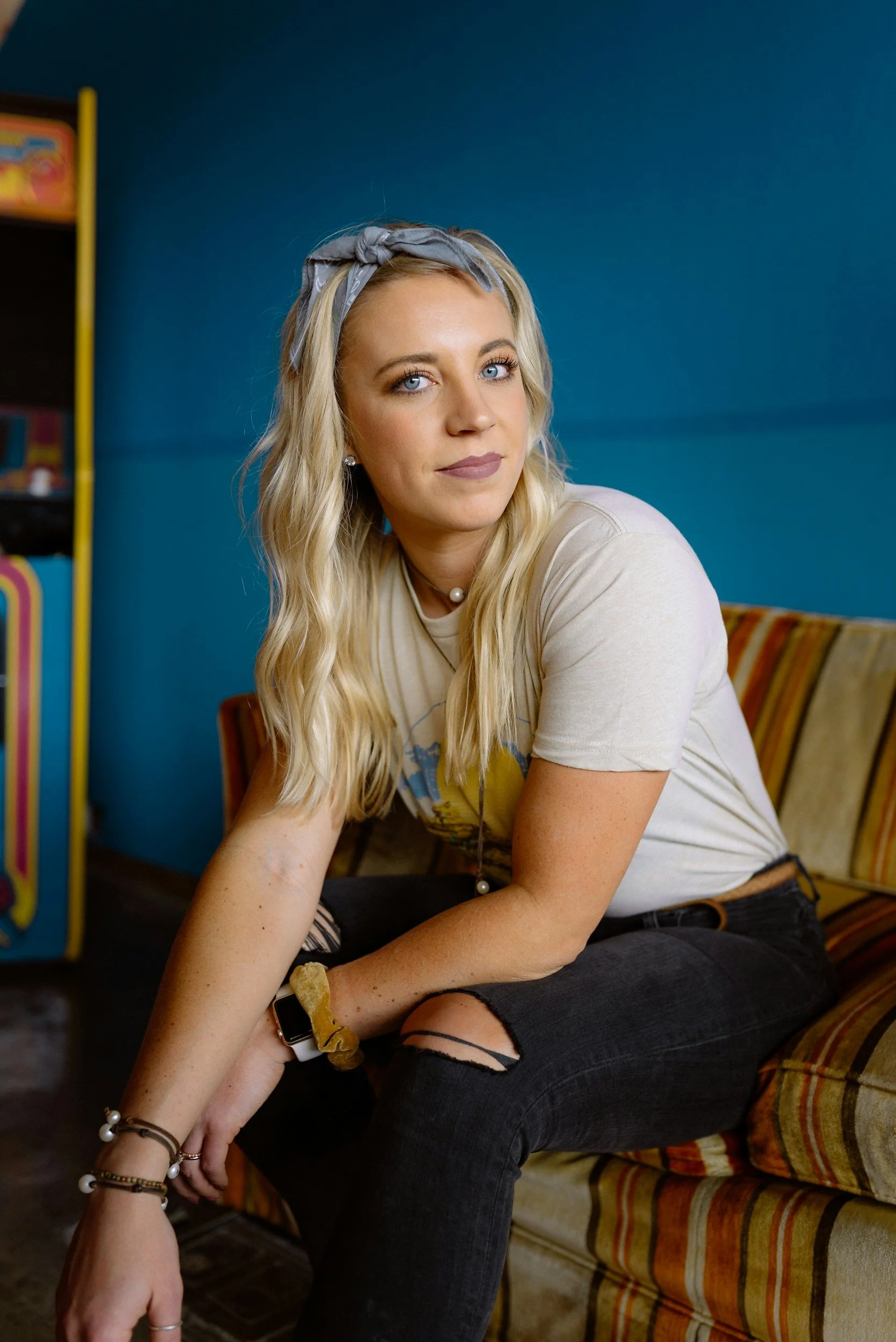 A young woman with long blonde wavy hair, light makeup, wearing a grey headband, pearl necklace, and a light-colored t-shirt, sitting on a vintage striped couch against a blue wall.