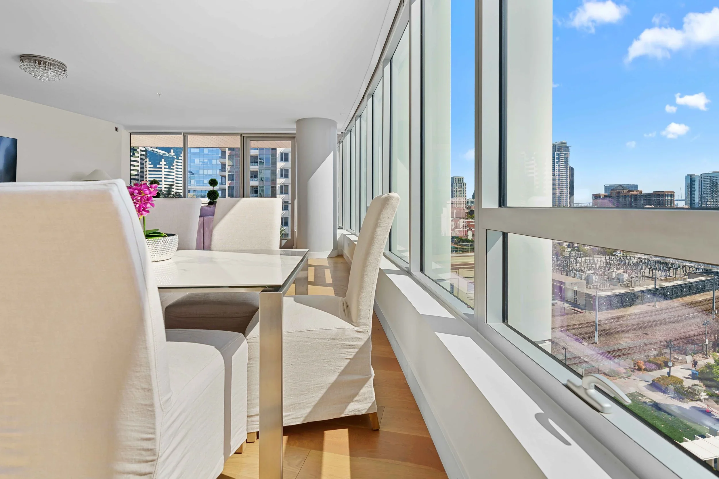 Modern dining area with white chairs, a orchid centerpiece on the table, and large floor-to-ceiling windows showing a cityscape with tall buildings.