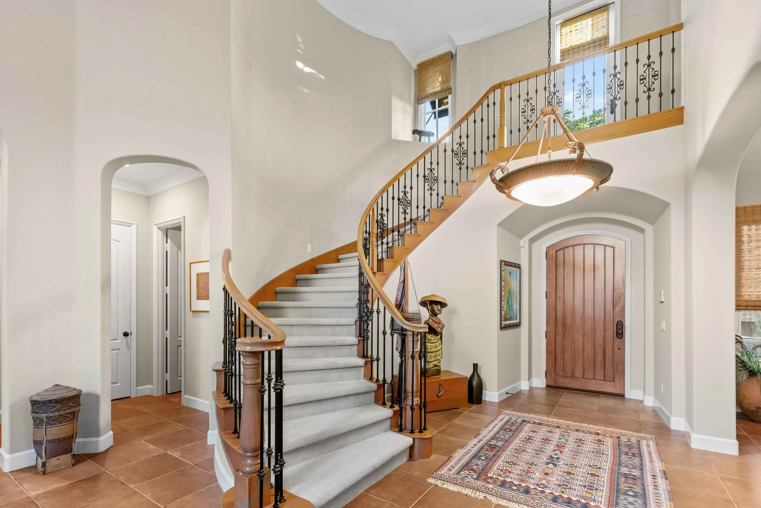 Entryway with a curved staircase, wooden handrail, and black decorative iron balusters. A tan front door with an arched top, a colorful patterned rug, and a chandelier hanging from the ceiling. There are a few pieces of artwork and decorative items, 