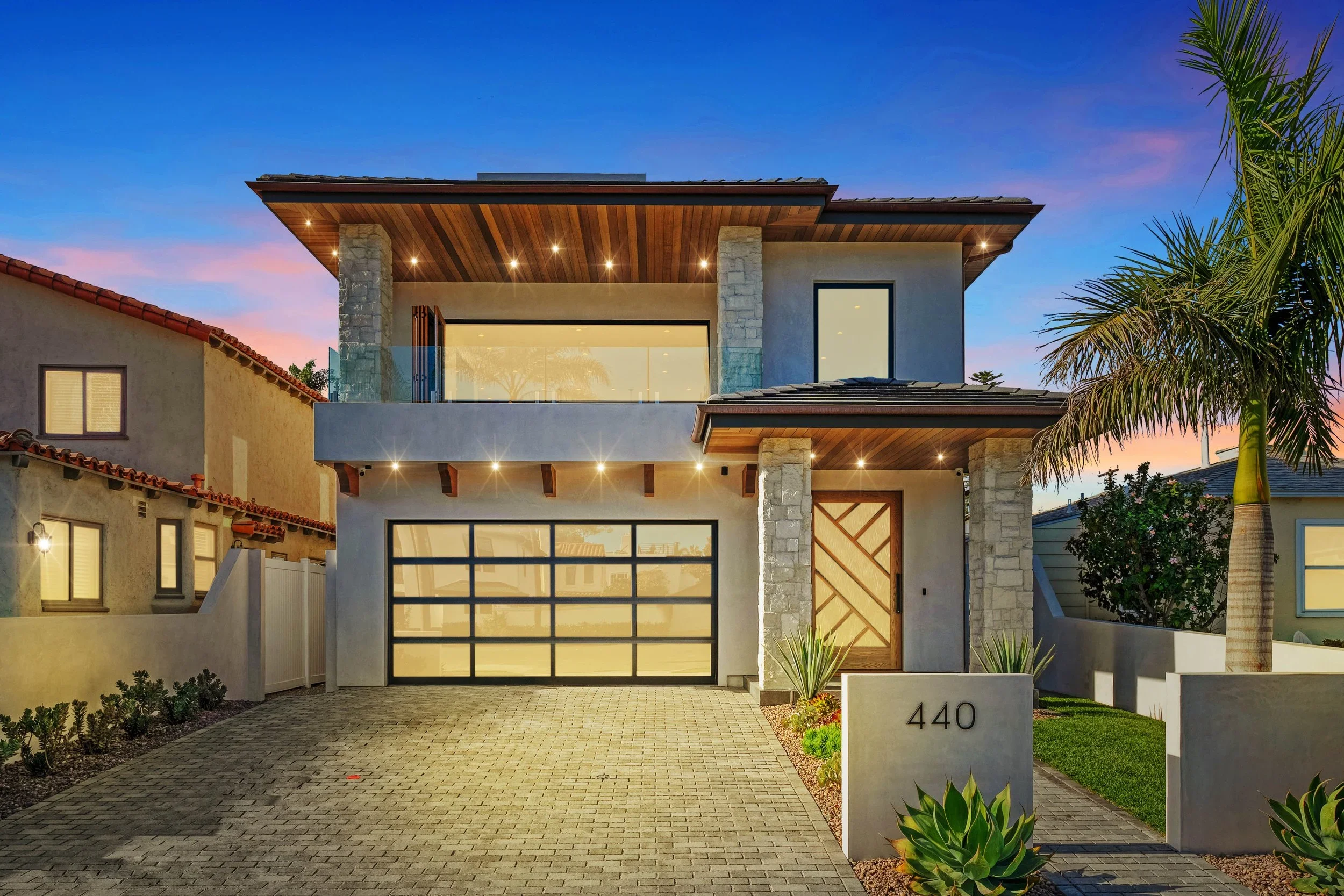 Modern two-story house with stone columns, a large glass garage door, and a wooden front door, situated in a tropical setting with palm trees, at dusk.