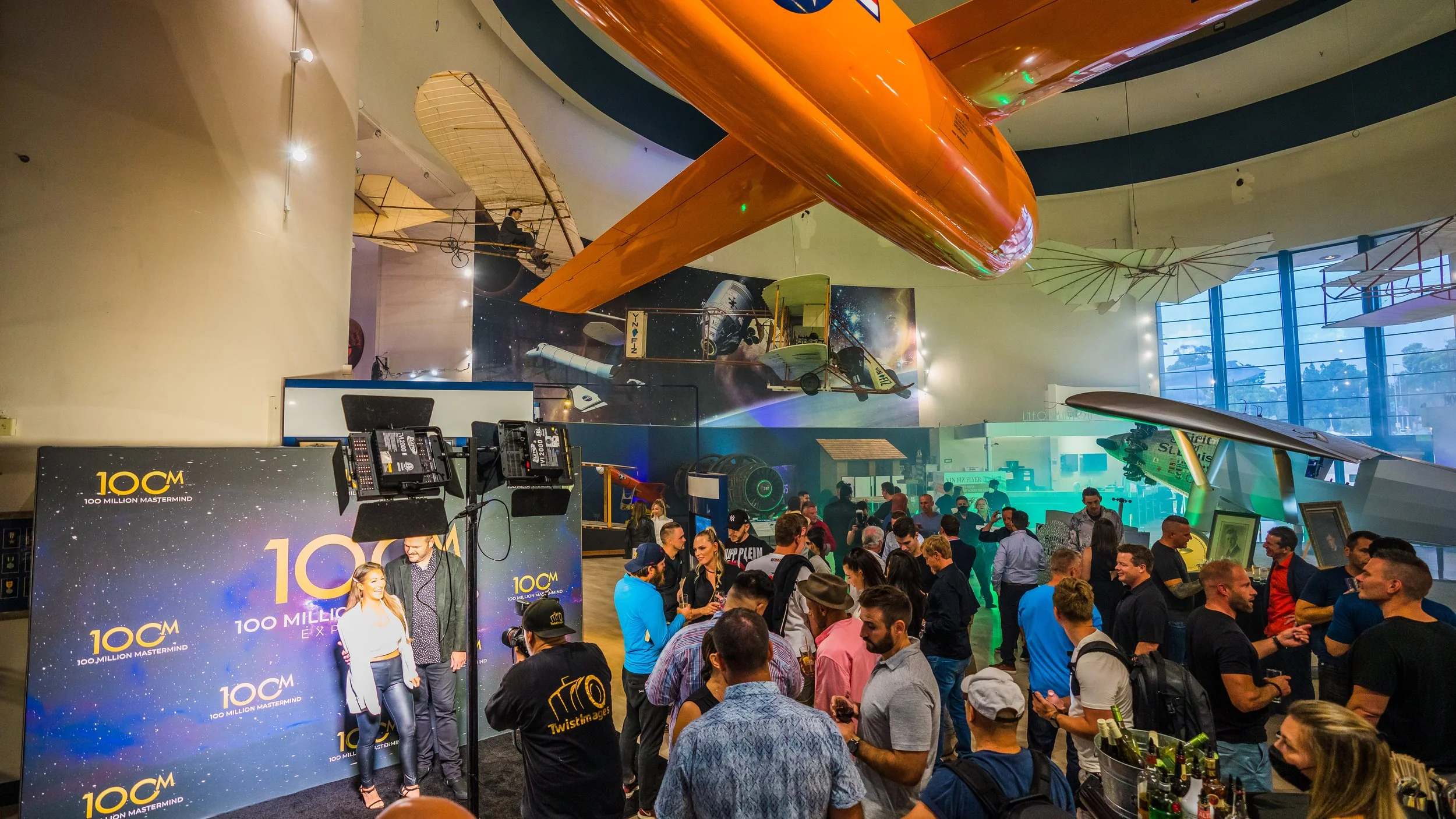A busy event space with a crowd of people gathered around a photo booth that has a backdrop with the '10CM 100 Million Mastermind' logo. The ceiling features large, vintage-style airplanes and space-themed decorations, including a mural of spacecraft