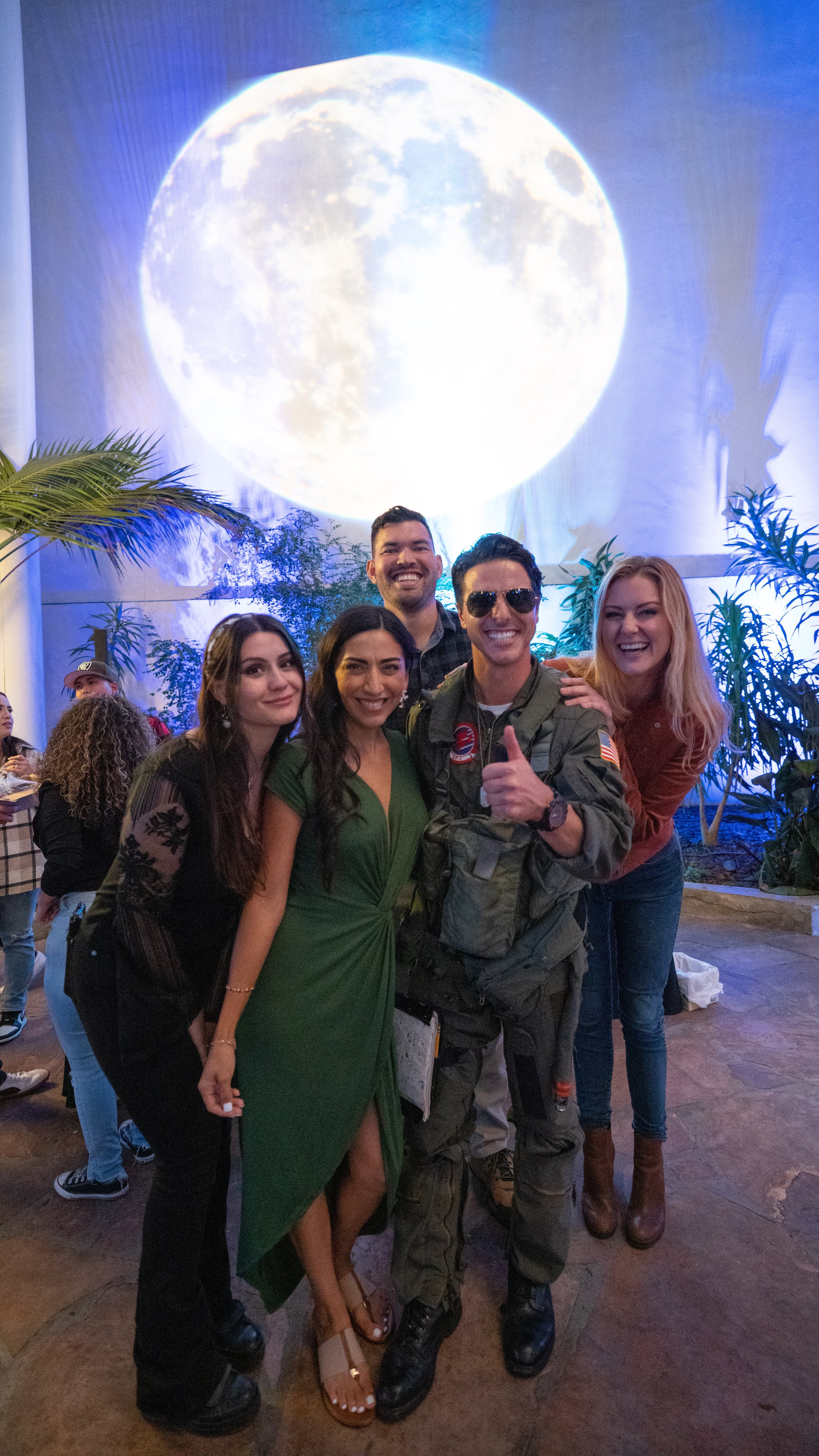 Group of five people smiling, posing together in front of a large projection of the moon, with plants around them.