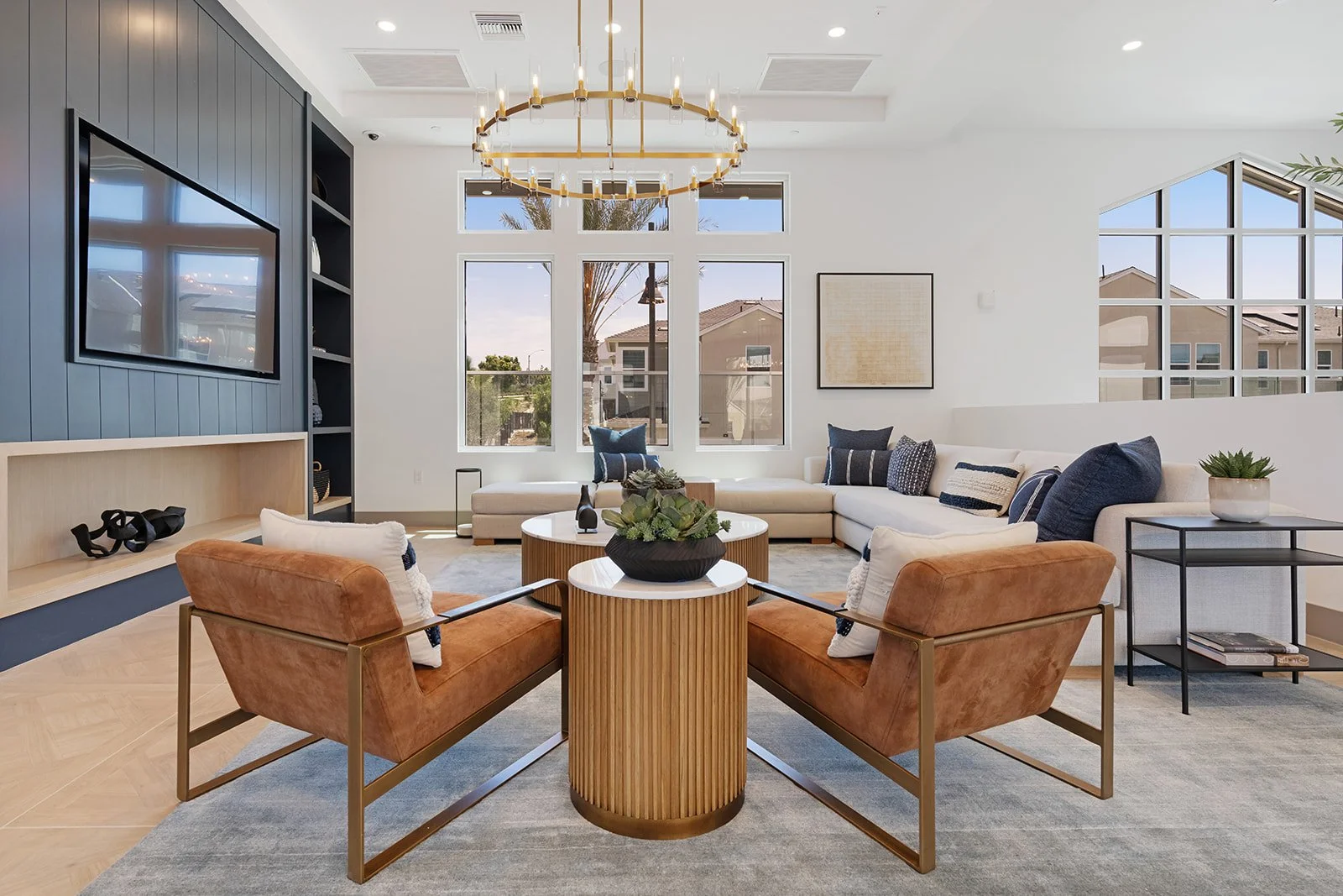 Modern living room with white walls, large windows, a black entertainment center with a mounted TV, a gold chandelier, an off-white sectional sofa, two orange armchairs, and decorative pillows, with a mix of natural light and minimalist decor.