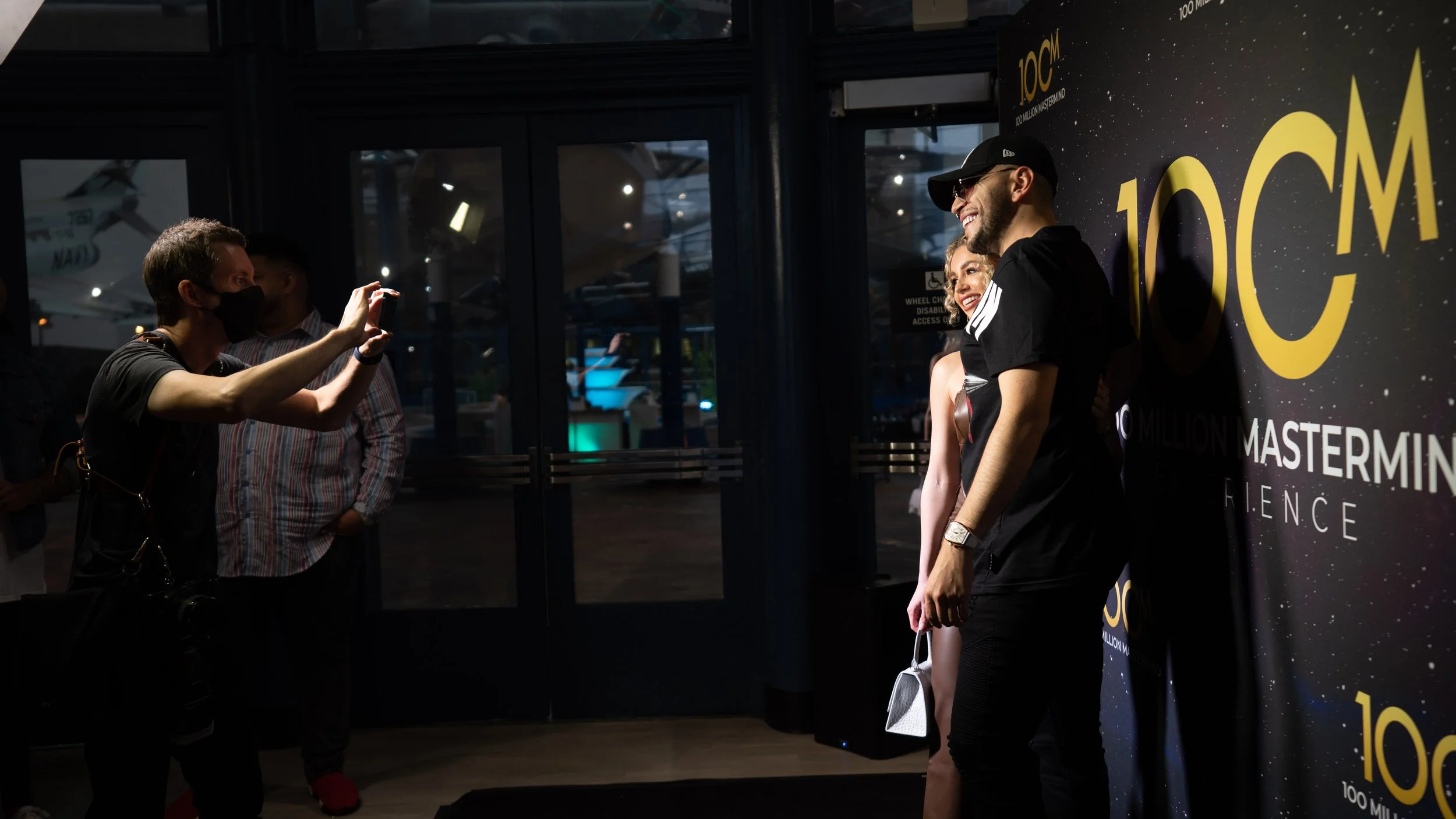 People taking a photo of a smiling man and woman at an event with a large backdrop that reads "100M" and "Mastermind." The setting appears to be indoors with dark lighting.