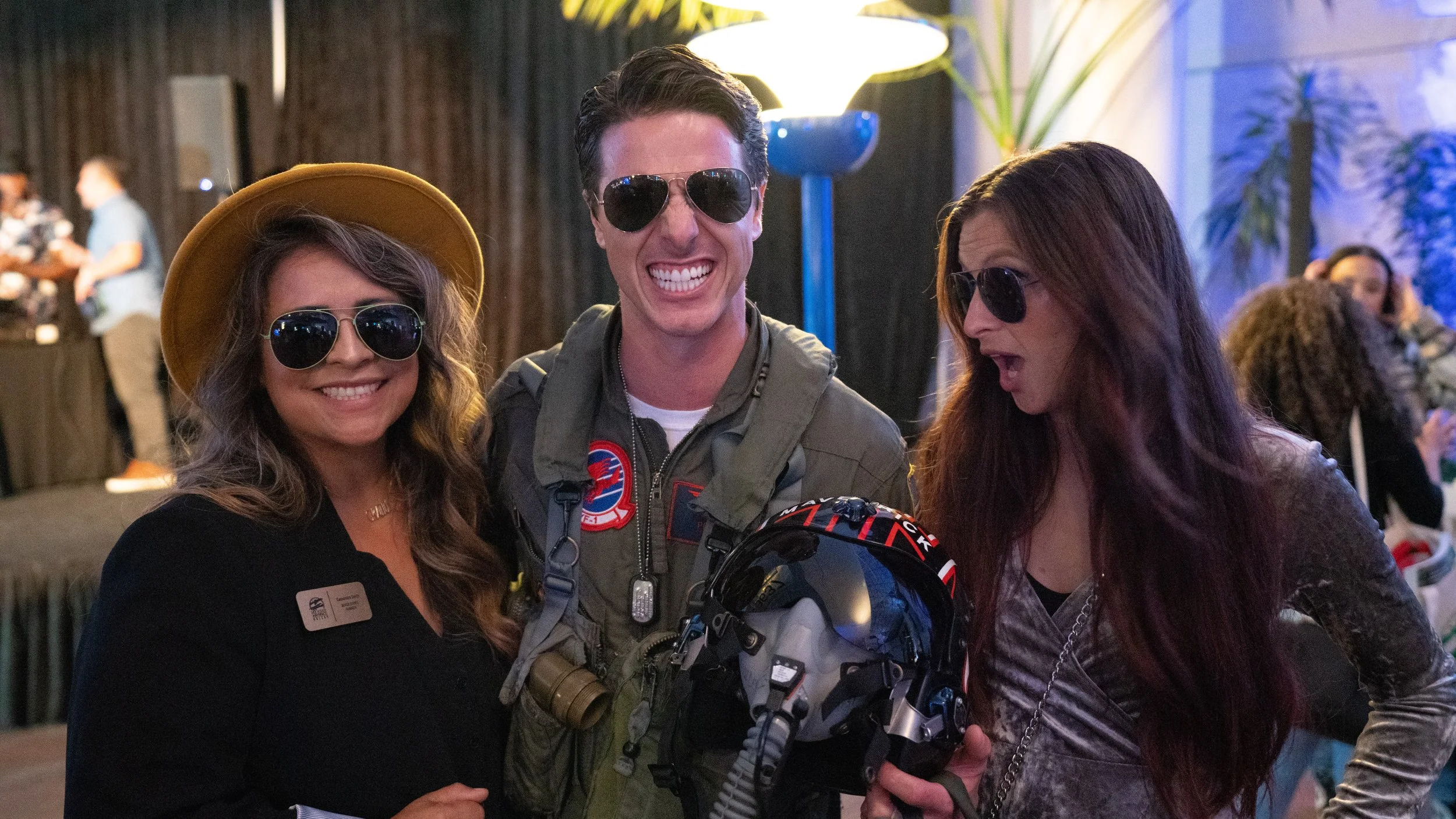 Three people wearing sunglasses, smiling, in a lively indoor setting. The woman on the left has long wavy hair, a yellow hat, and a name tag. The man in the middle is dressed like an astronaut with a flight suit and holding a helmet. The woman on the