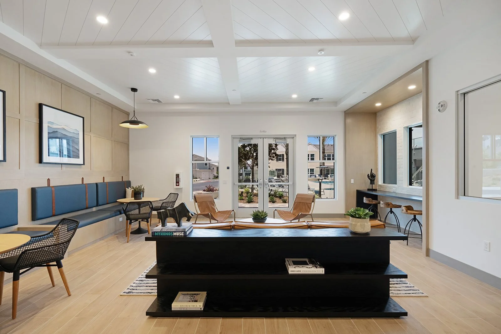 An airy, modern lounge area with light-colored wooden flooring, white walls, and large windows opening to an outdoor view. There are various seating options including a long blue cushioned bench, black mesh chairs around small round tables, and two t