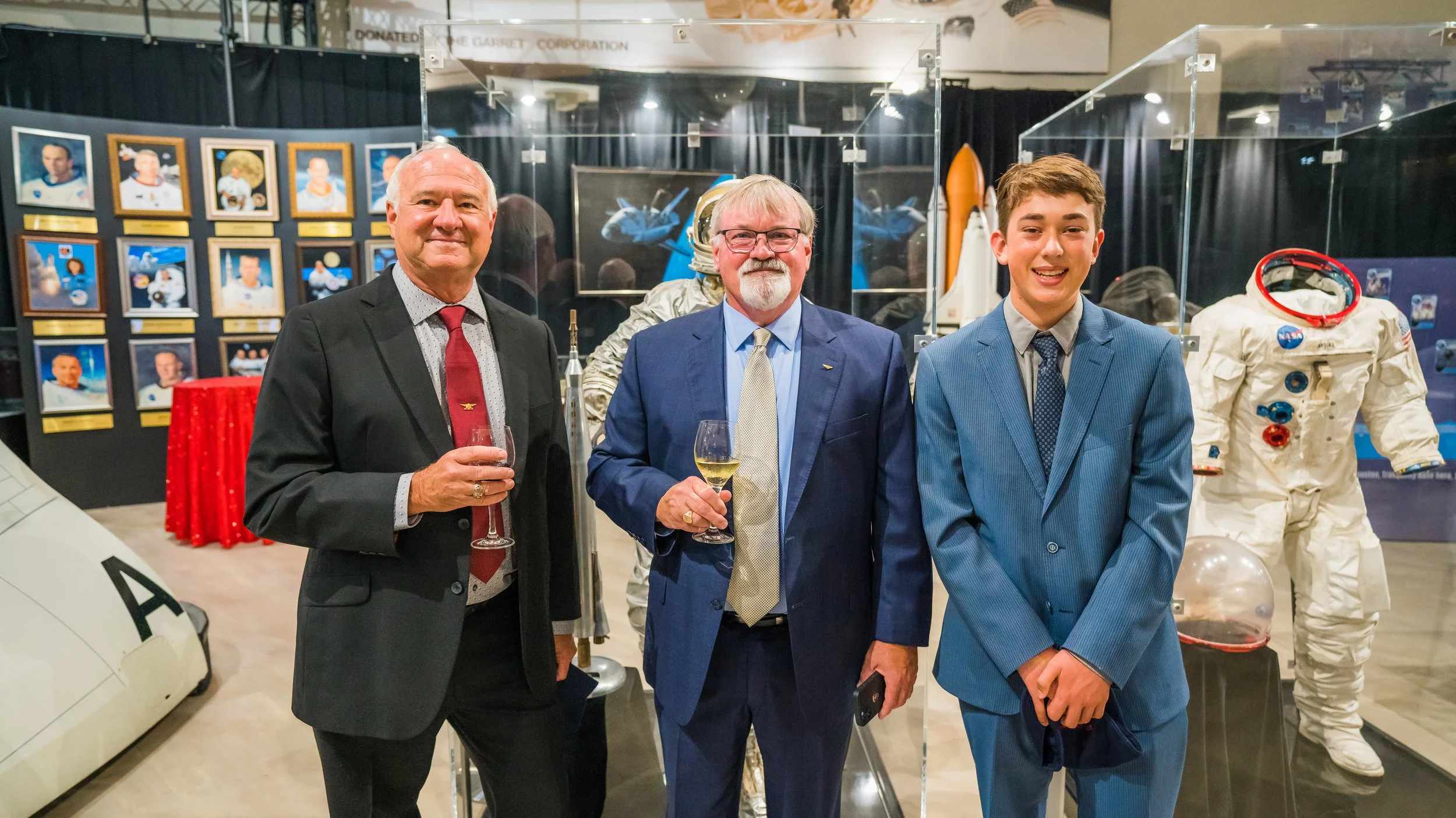 Three men in suits holding glasses of wine posing for a photo at a space museum with framed astronaut photos and space suits in the background.
