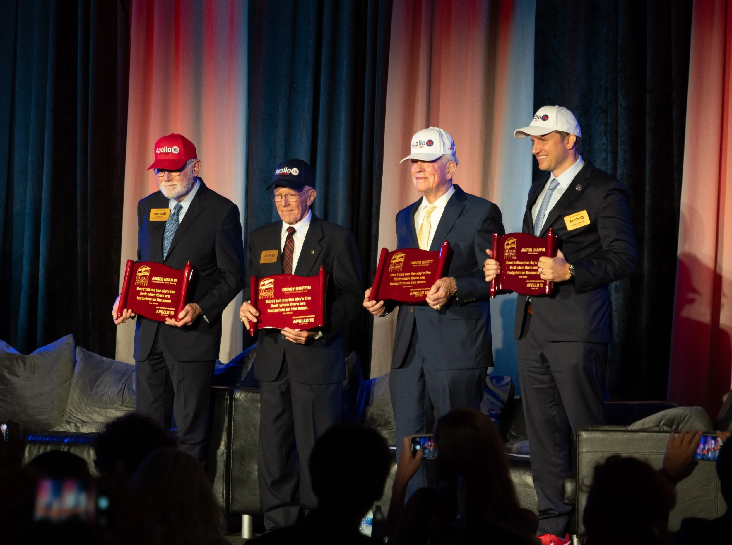 Four men in suits holding red plaques, standing in front of a stage curtain, with some audience members taking photos.