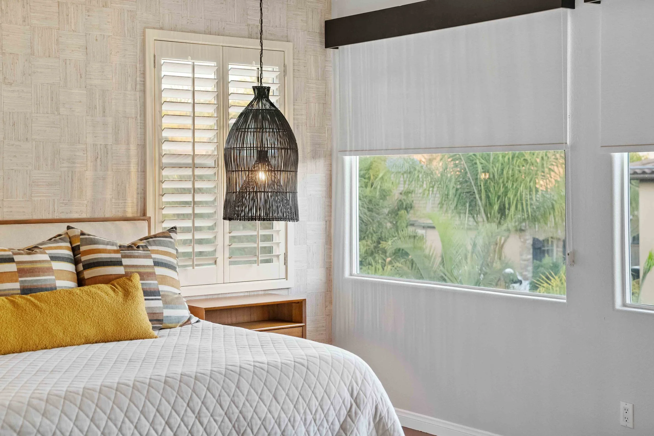 Bedroom with a bed, striped pillows, a yellow pillow, a wooden nightstand, white window shutters, black hanging pendant lamp, large window with rolled white blinds, and greenery outside.