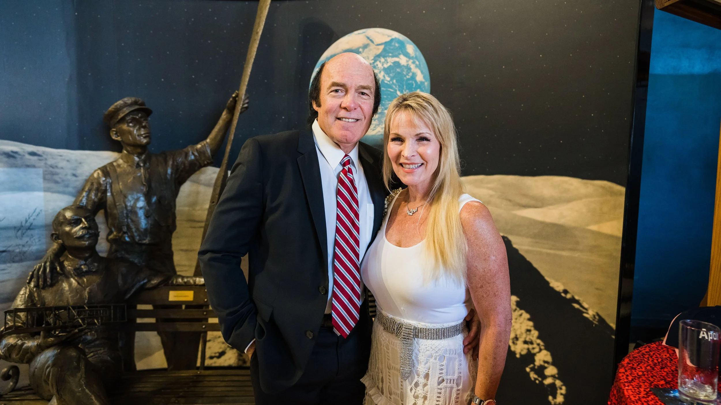 A man and woman standing together indoors, smiling for the camera, with a space-themed backdrop featuring an astronaut statue, the Earth, and the Moon.