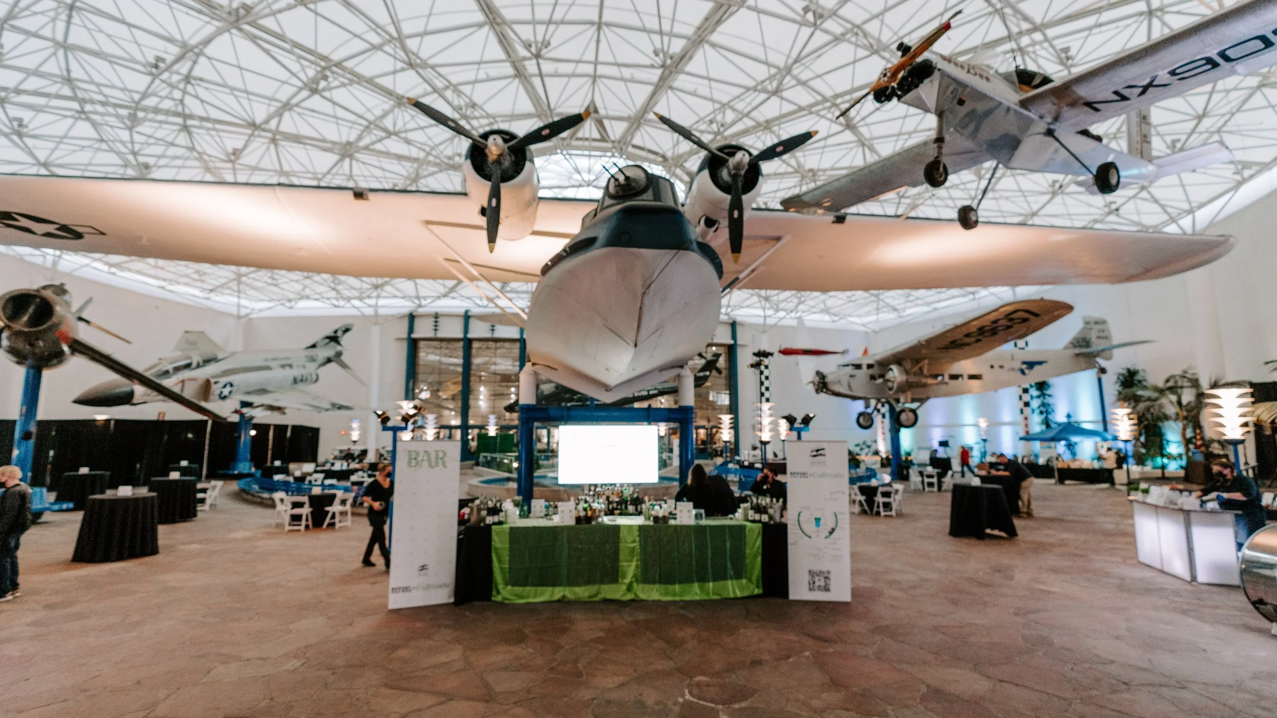 Indoor museum or exhibition with suspended aircraft, including a large seaplane with pontoons and smaller jets, seating areas, and people walking around.