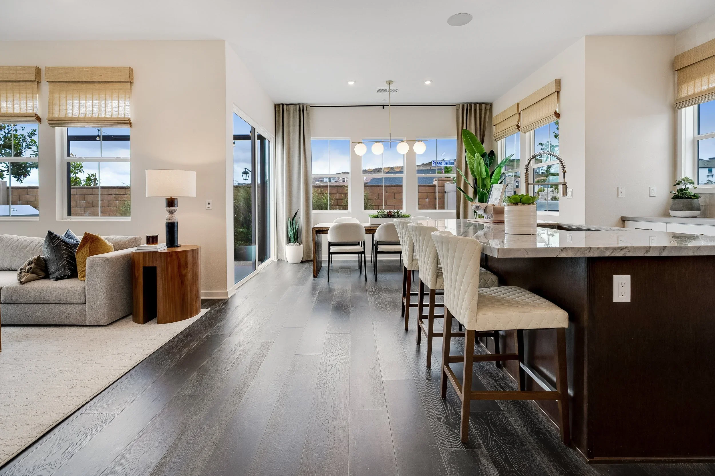 Open-concept living and dining area with large windows, wooden flooring, a kitchen island with barstools, a dining table with chairs, a sofa with cushions, and potted plants.