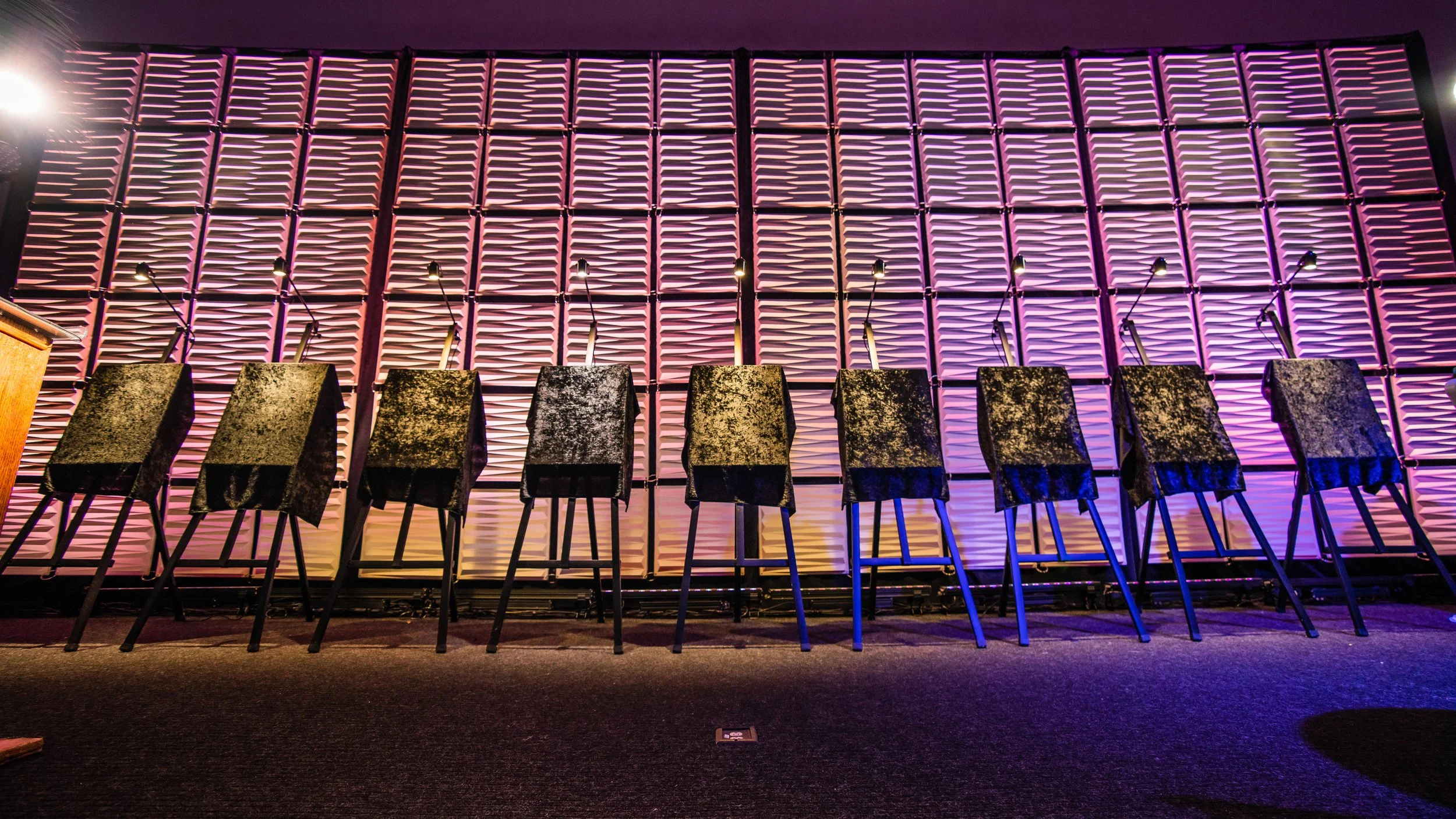 Eight chairs covered with black cloths and standing behind a panel decorated with textured pink and purple lighting.