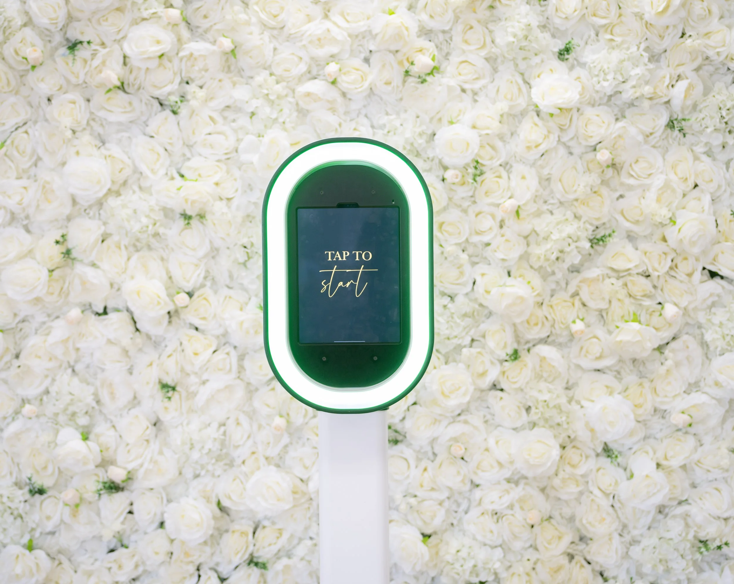 A modern electronic kiosk with a touchscreen displaying 'TAP TO start,' placed against a background of white roses and florals.