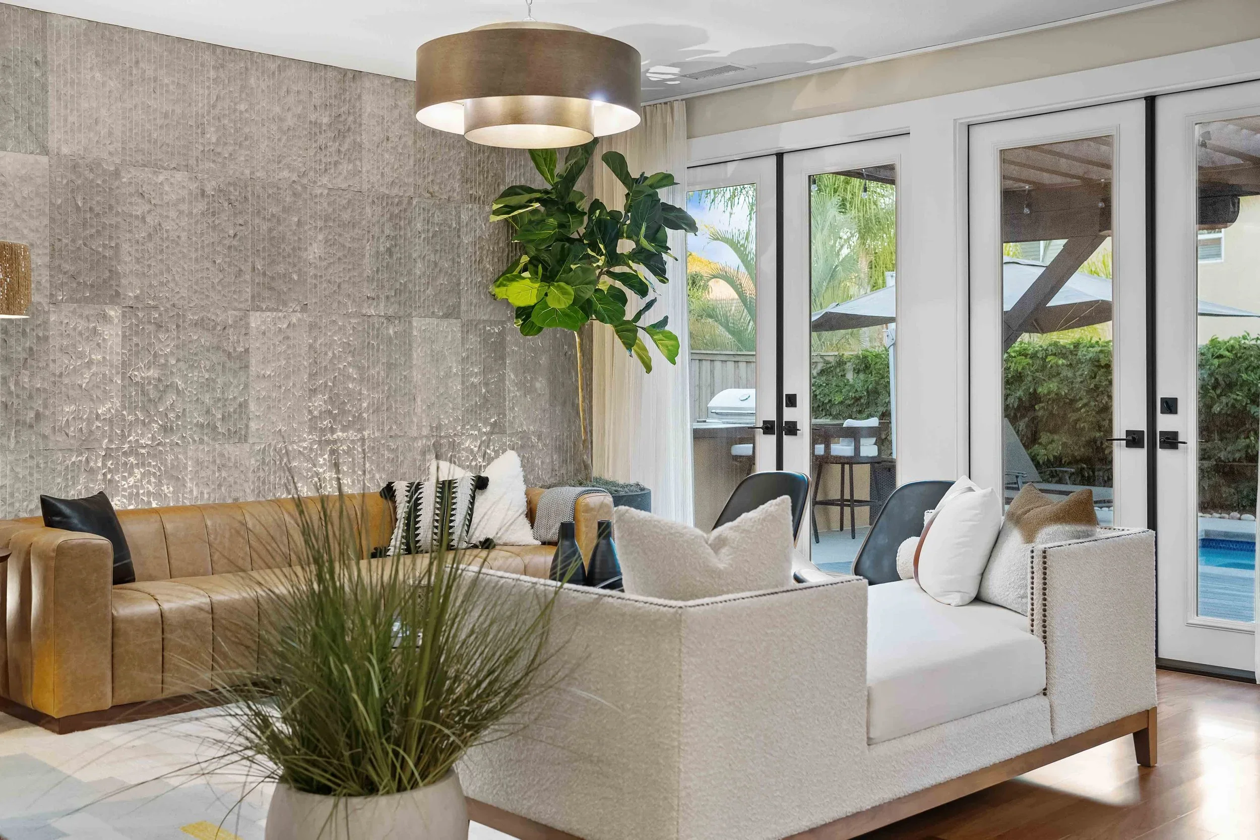 Living room with beige and white furniture, large sliding glass doors leading to an outdoor patio with a grill and umbrellas, potted plants, and modern décor.