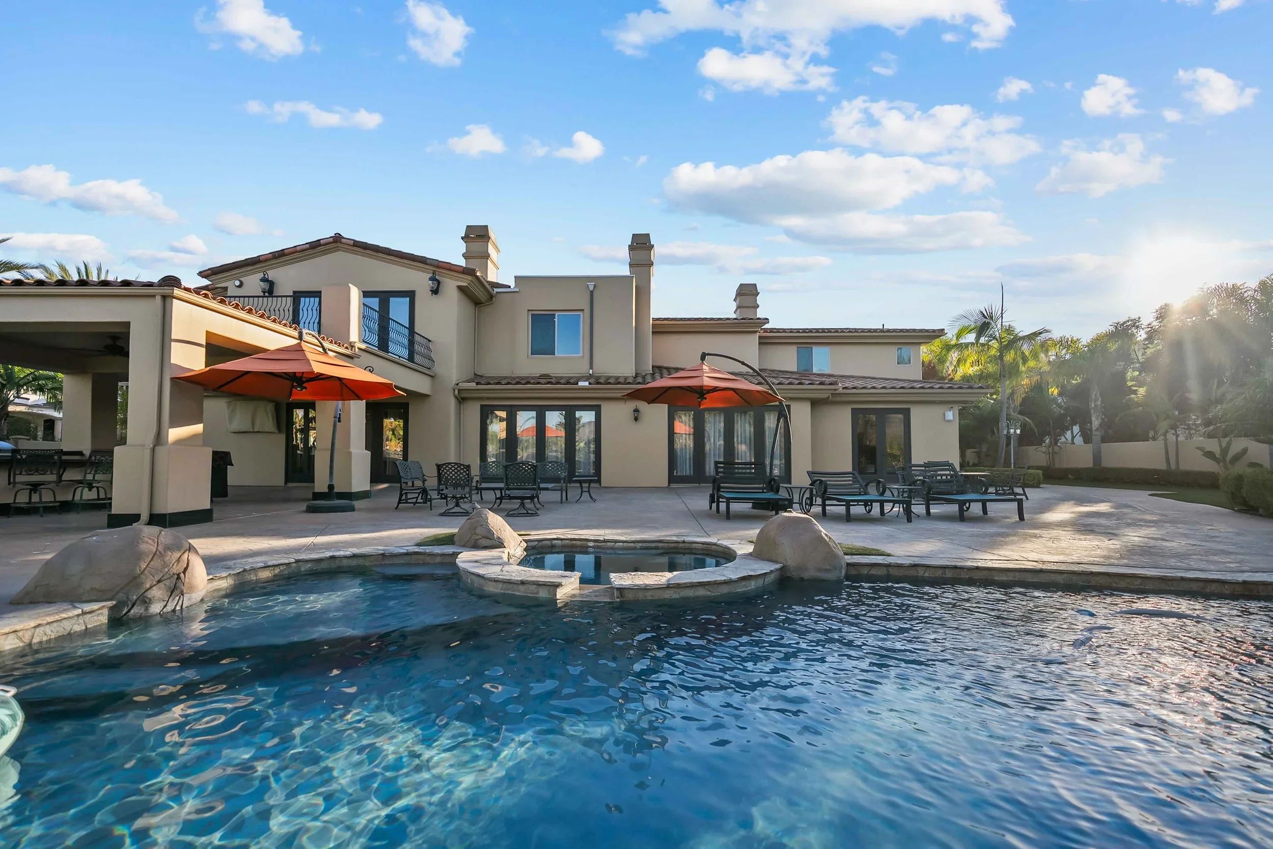 Large residential house with a backyard pool surrounded by patio furniture and umbrellas, with trees and a partly cloudy sky in the background.