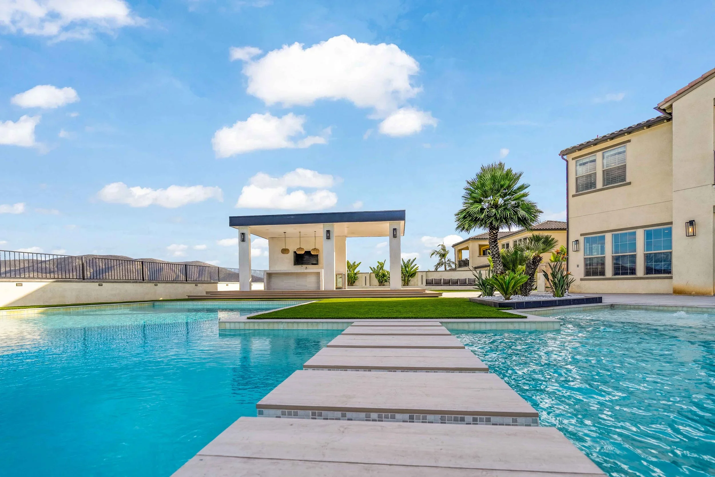 Luxury backyard with swimming pool, stepping stones, palm trees, a gazebo with outdoor tv and seating, modern house with large windows, blue sky with clouds.