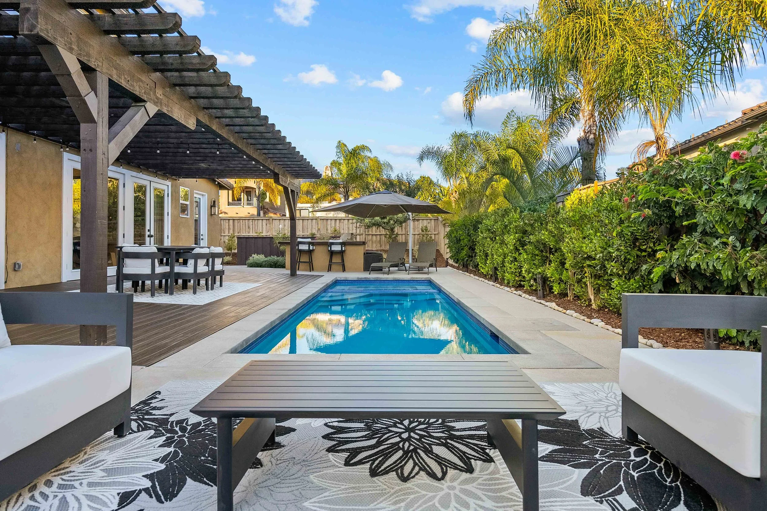 Backyard with rectangular pool, outdoor seating area, bar, and lounge chairs, surrounded by green bushes and palm trees under a blue sky.