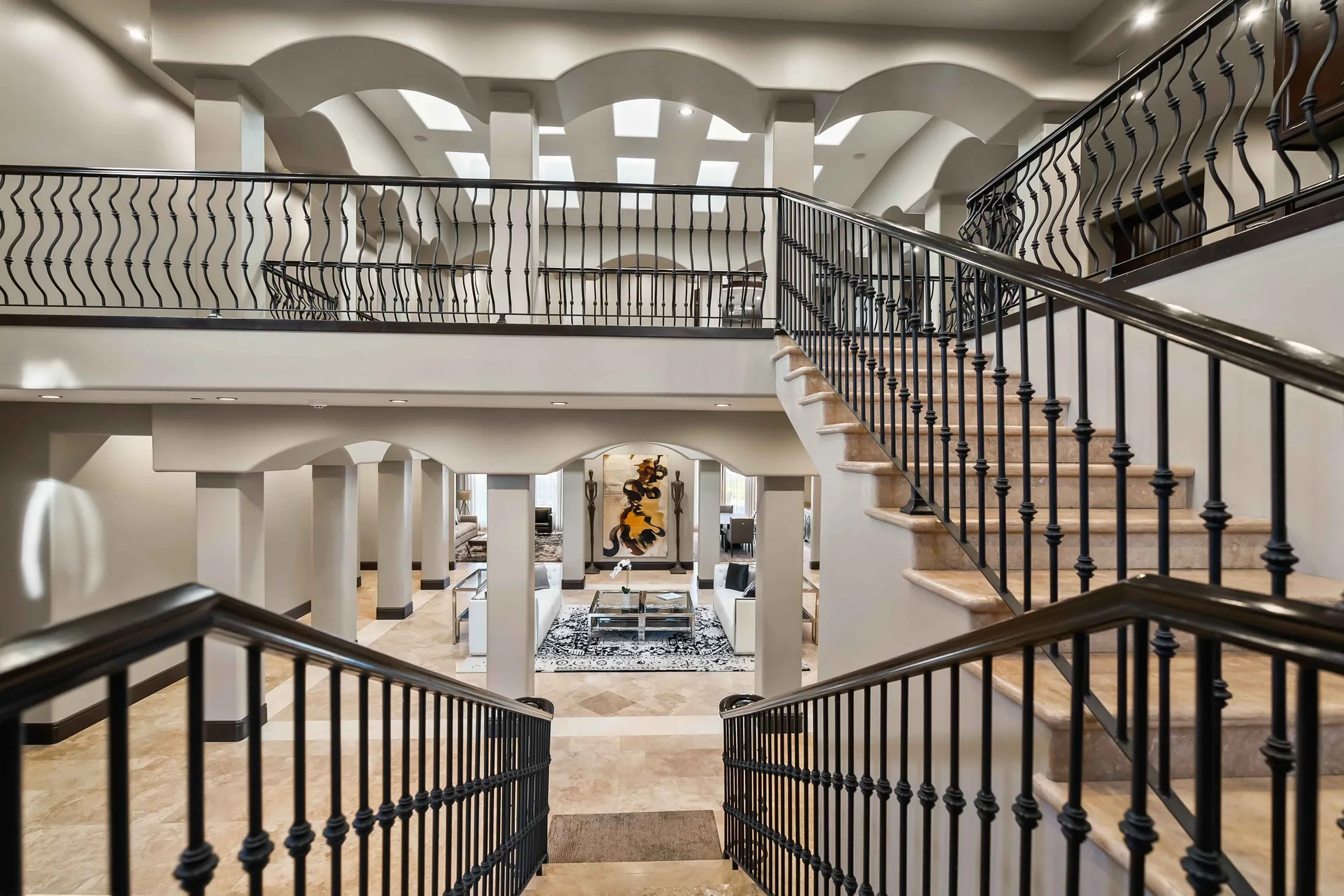 Interior of a spacious, multi-level house with staircase and balcony, featuring black railings, white walls, and modern furniture in the living area.