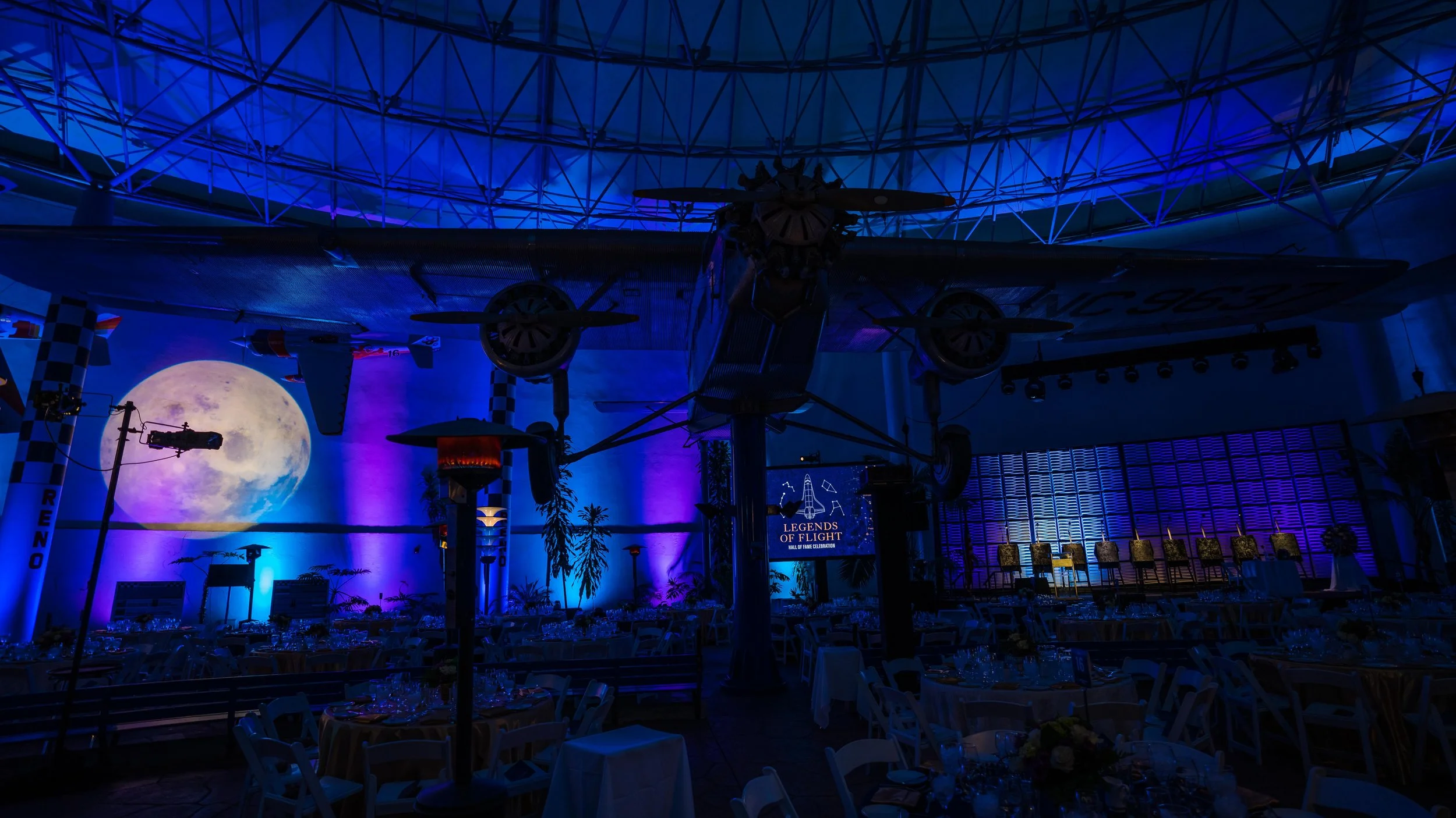 Indoor event space decorated with aviation theme, featuring a large model airplane hanging from the ceiling, a projection of the moon on the wall, tables set with glassware and floral centerpieces, and a sign that reads 'Legends of Flight'.