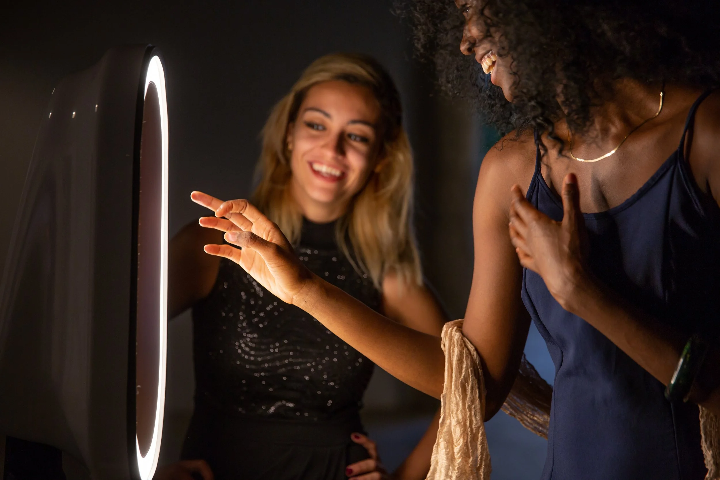 Two women smiling and interacting with a glowing ring-shaped object in a dark room.