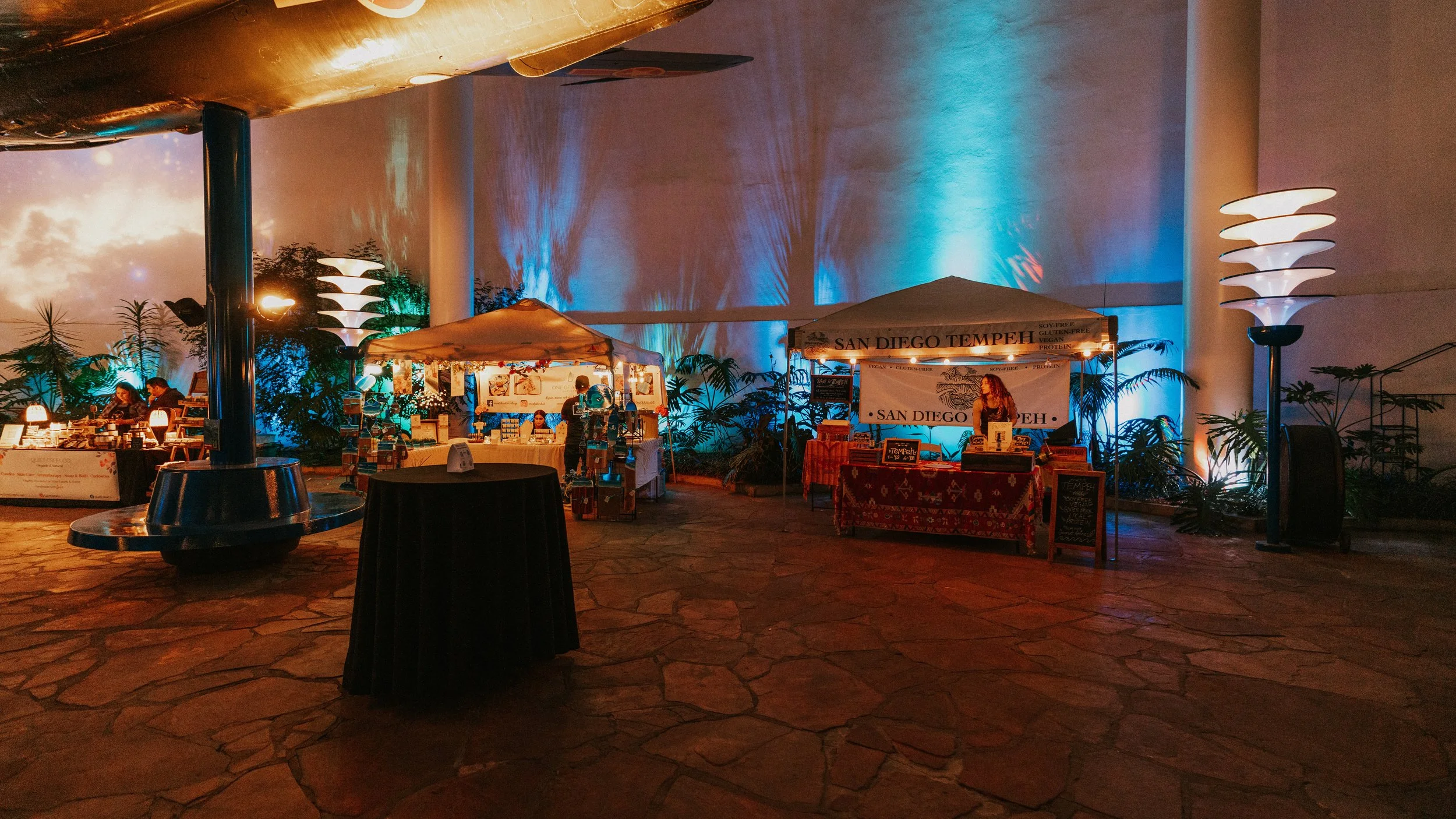 Indoor event space with vendor booths, tables, and decorative lighting, including illuminated plants and a large wall with blue and orange lighting effects.