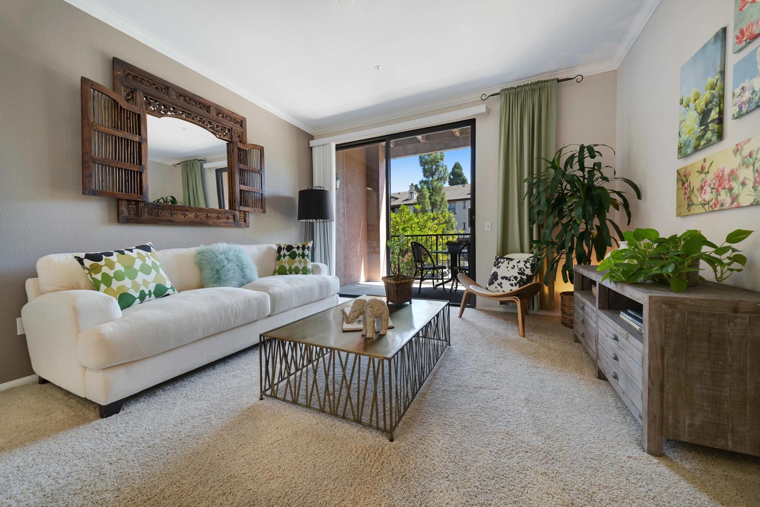 Living room with white sofa, decorative pillows, wooden wall mirror, black floor lamp, glass sliding door leading to a balcony with outdoor furniture, potted plants, a wooden sideboard, colorful paintings, and a spacious layout with natural light.