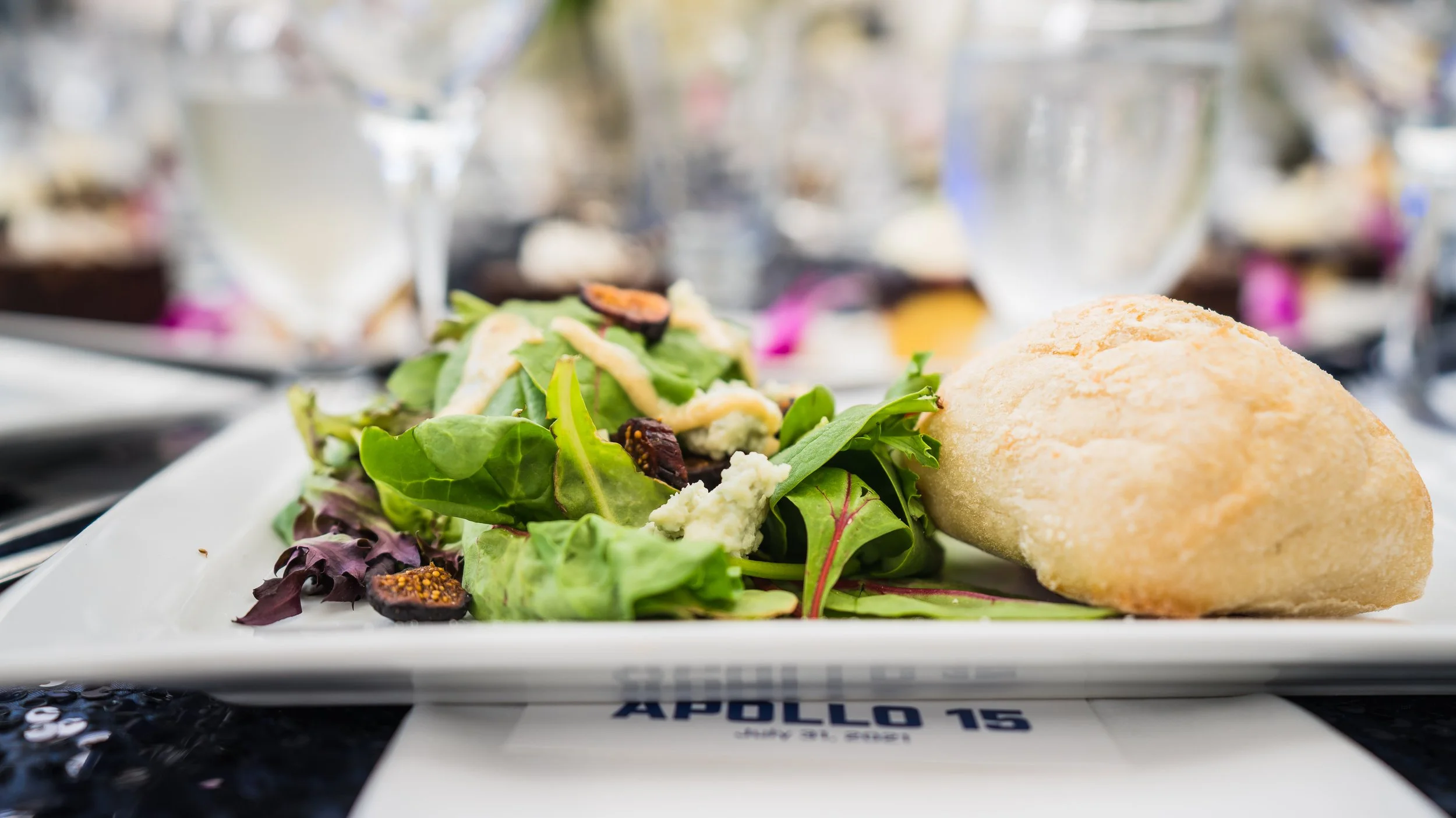 Salad with mixed greens, blue cheese crumbles, dried figs, and dressing, served alongside a bread roll on a white rectangular plate.