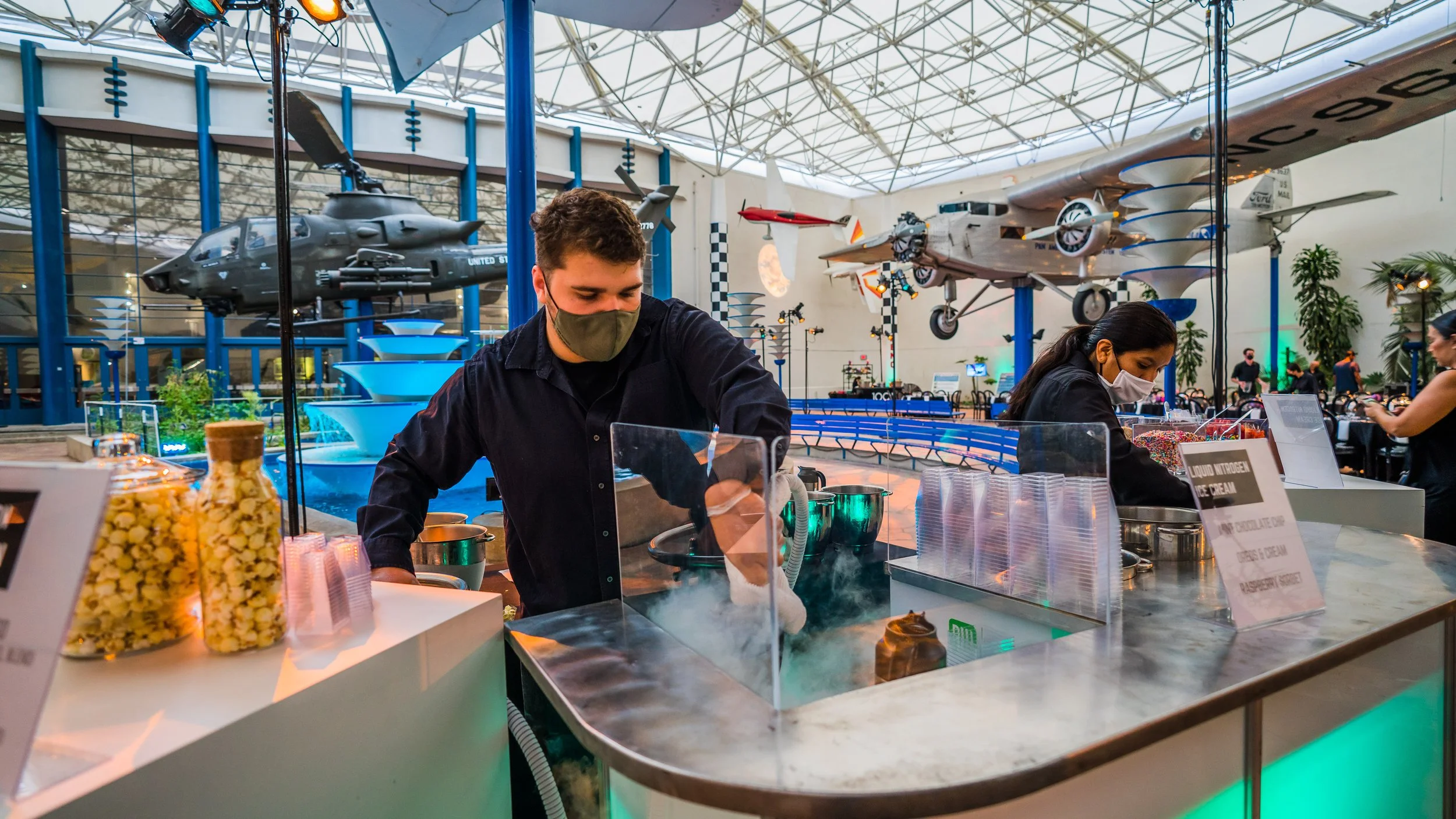 Two people wearing masks preparing ice cream at a dessert stand inside a large museum with airplanes suspended from the ceiling and a fountain in the background.