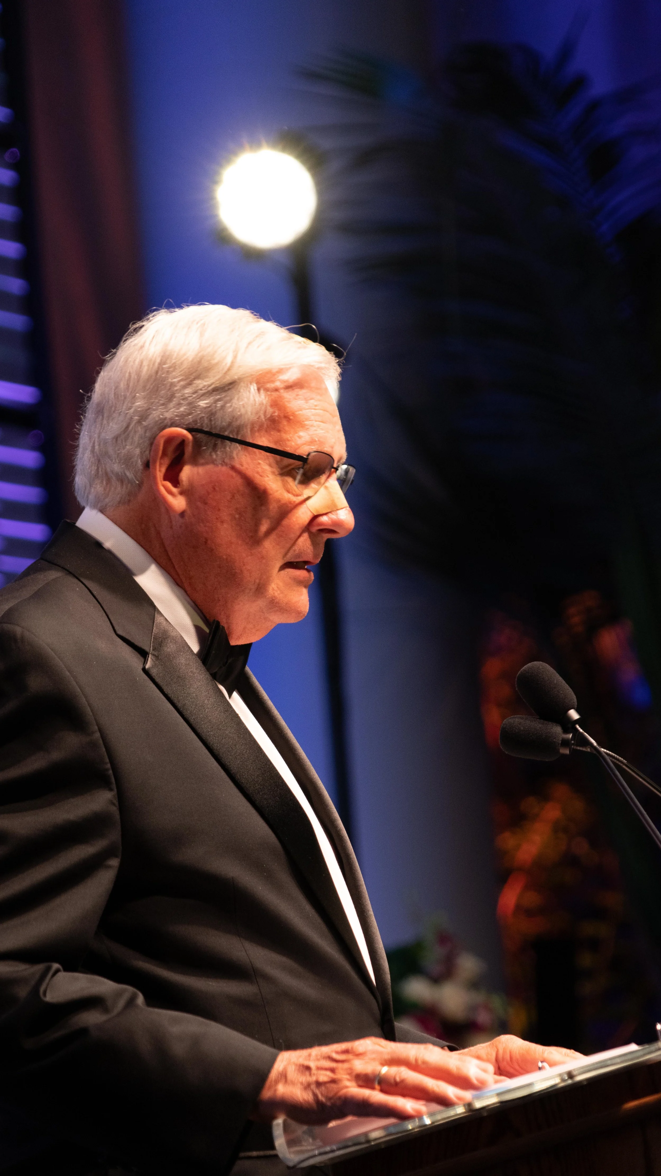 An elderly man in a black tuxedo with a bow tie, wearing glasses, speaking at a podium with microphones in a formal setting, with dim lighting and large green leaves in the background.