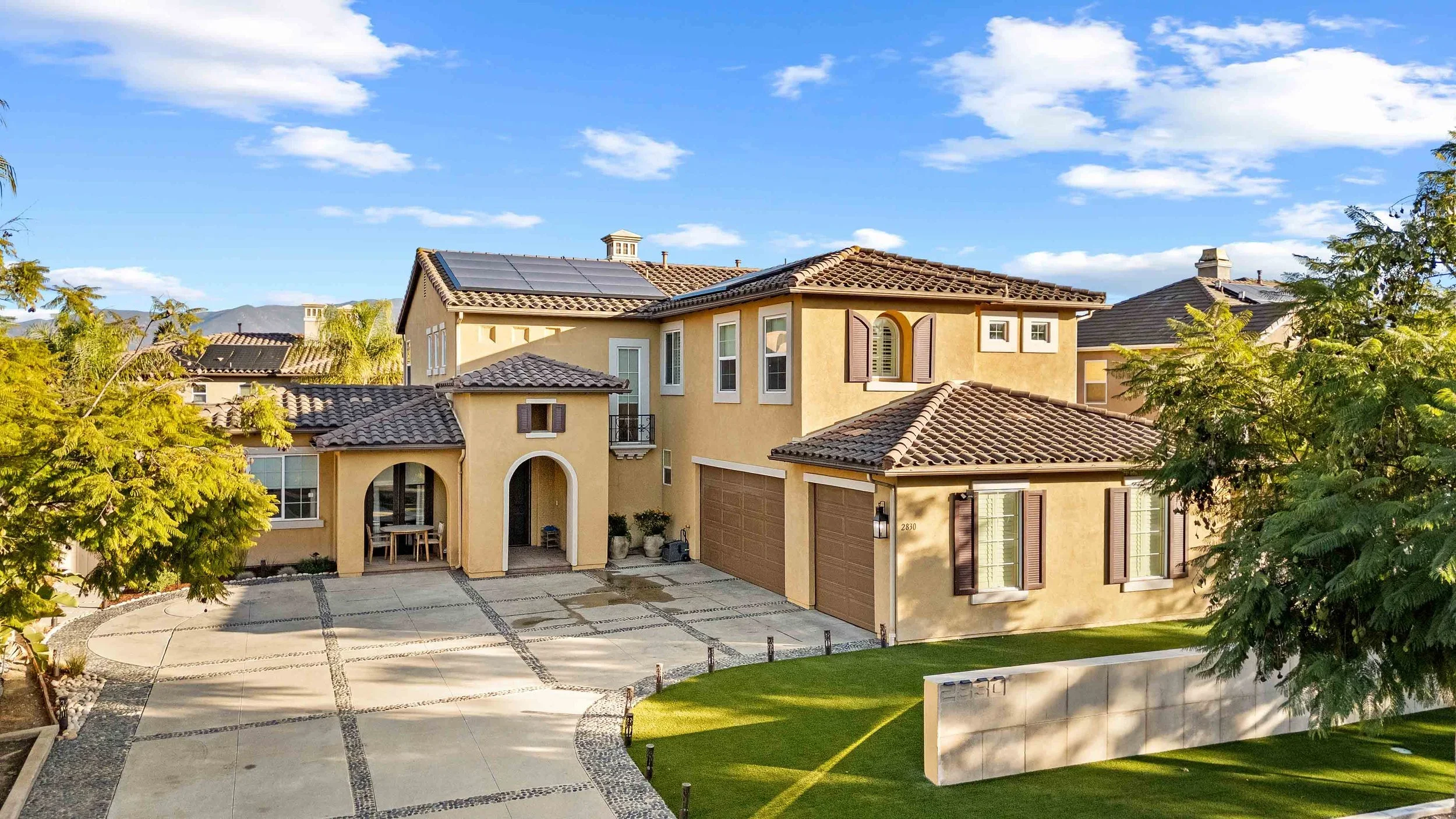Front view of a large two-story house with a tiled roof, surrounded by a well-maintained yard and trees, under a blue sky with clouds.