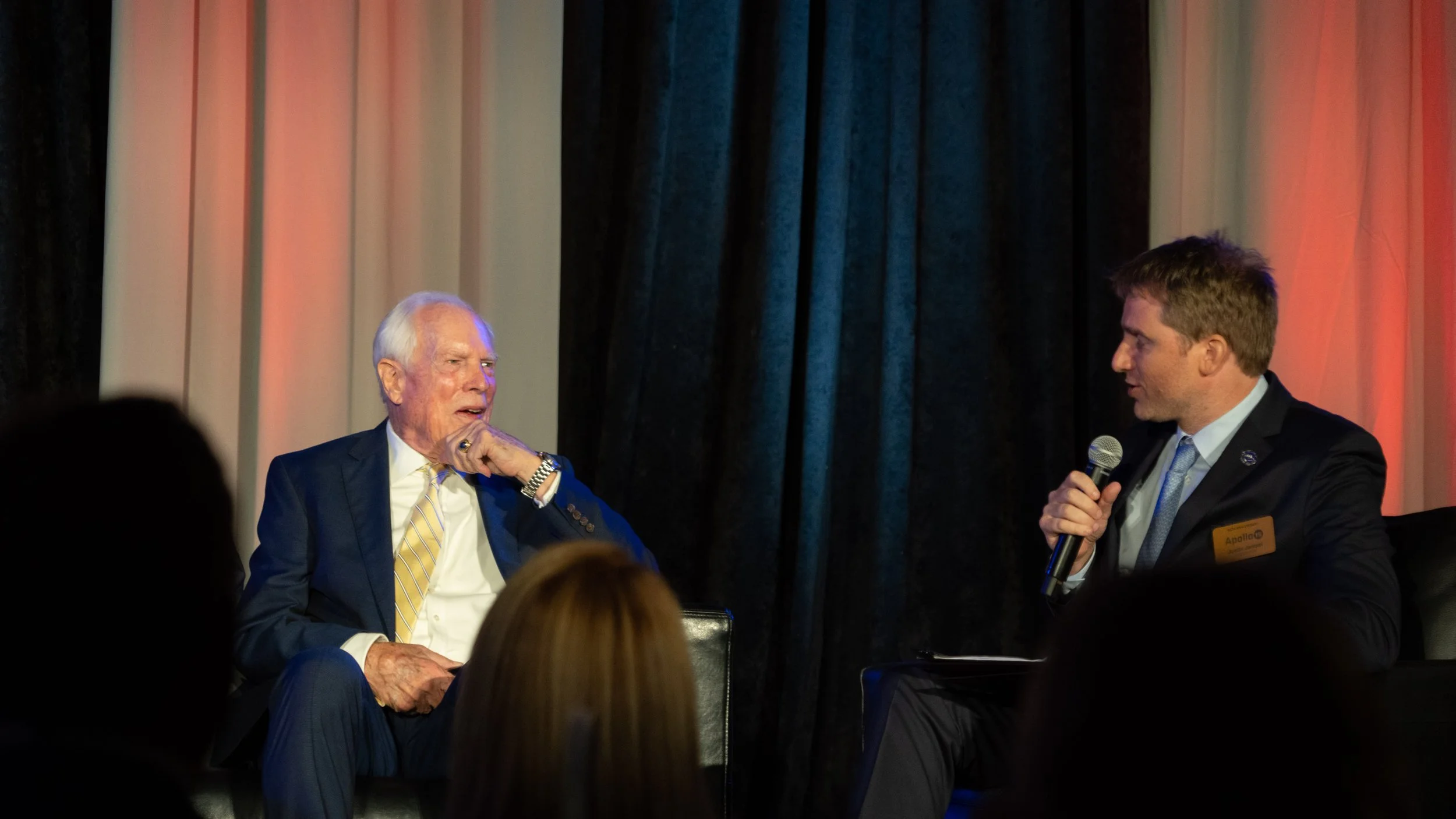 An older man and a younger man are sitting on stage in conversation, with the older man on the left, dressed in a dark suit and yellow striped tie, and the younger man on the right, in a dark suit, holding a microphone. Behind them are colorful curta