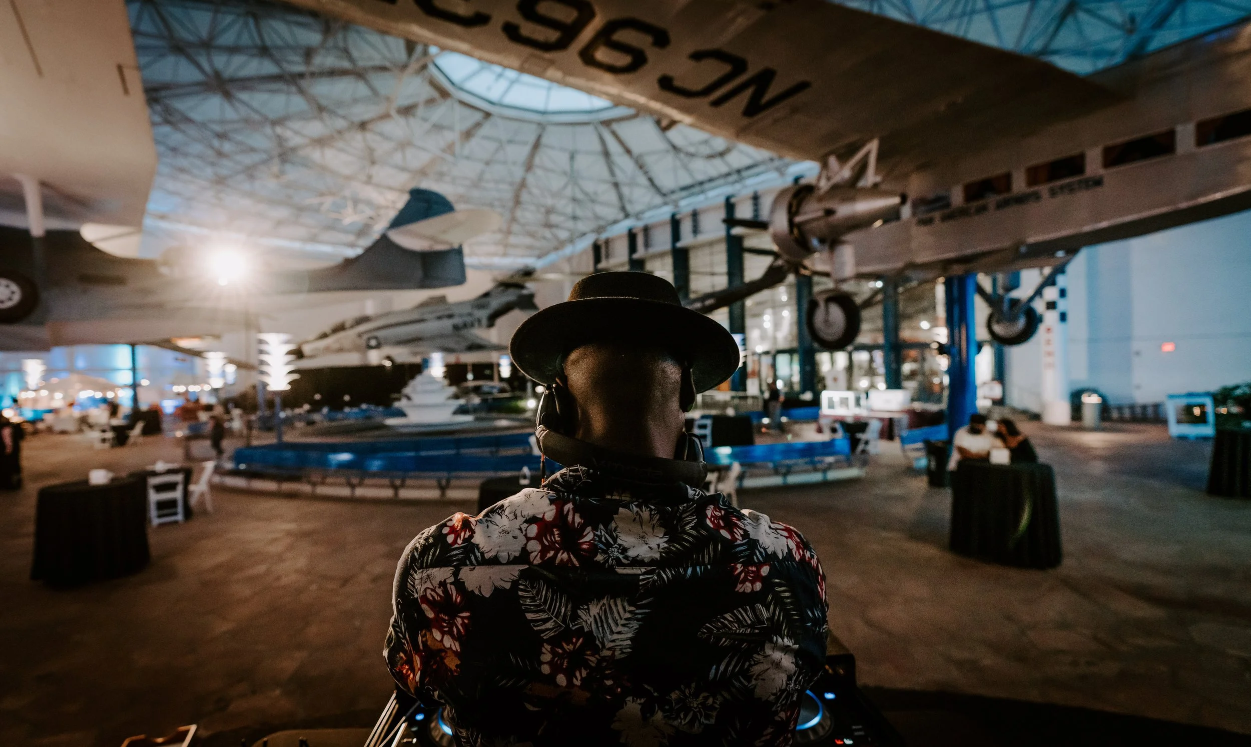 Person wearing a hat and floral shirt, facing an indoor aerospace museum with aircraft displays, including spaceships and planes, hanging from the ceiling.