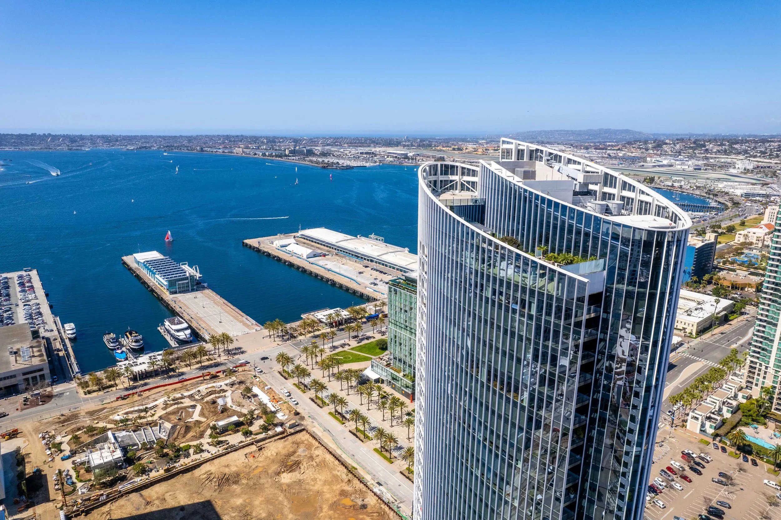 Aerial view of a modern urban waterfront with a tall, curved glass skyscraper, marina with boats, and cityscape in San Diego, California.