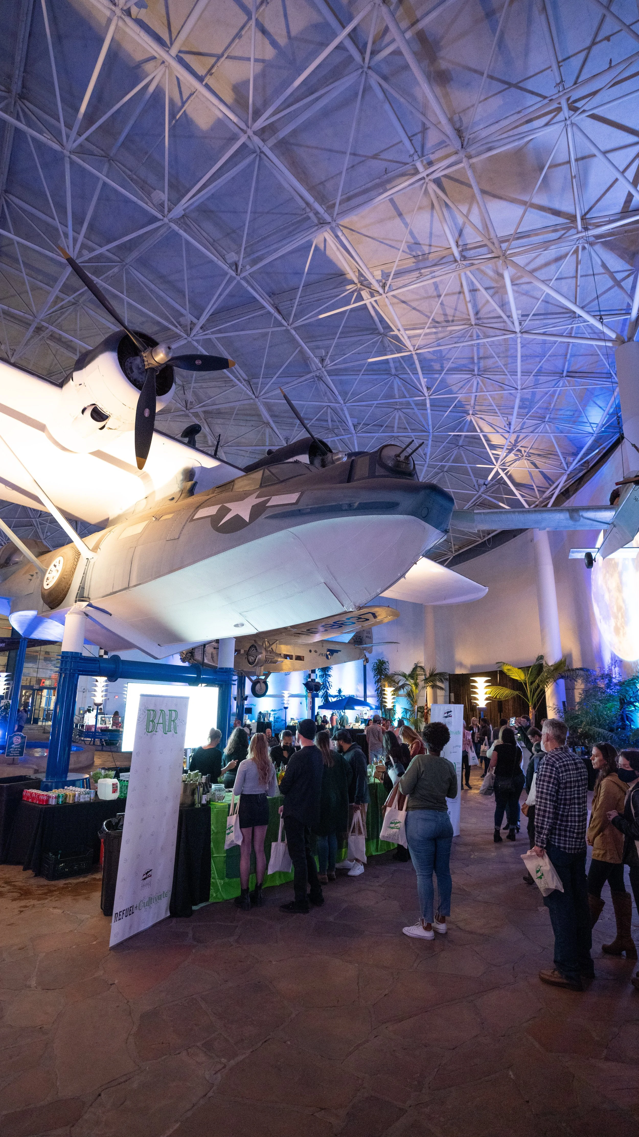 An indoor museum or exhibit hall with a large vintage airplane suspended from the ceiling, surrounded by people. There is a bar and various booths set up, with some donors or visitors holding bags. The ceiling has a geometric metal framework, and the
