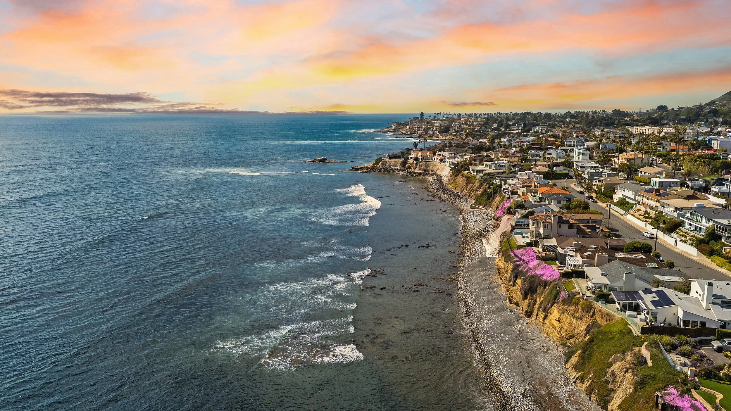 A coastal seaside neighborhood with homes on cliffs overlooking the ocean, during sunset with pink and orange clouds.