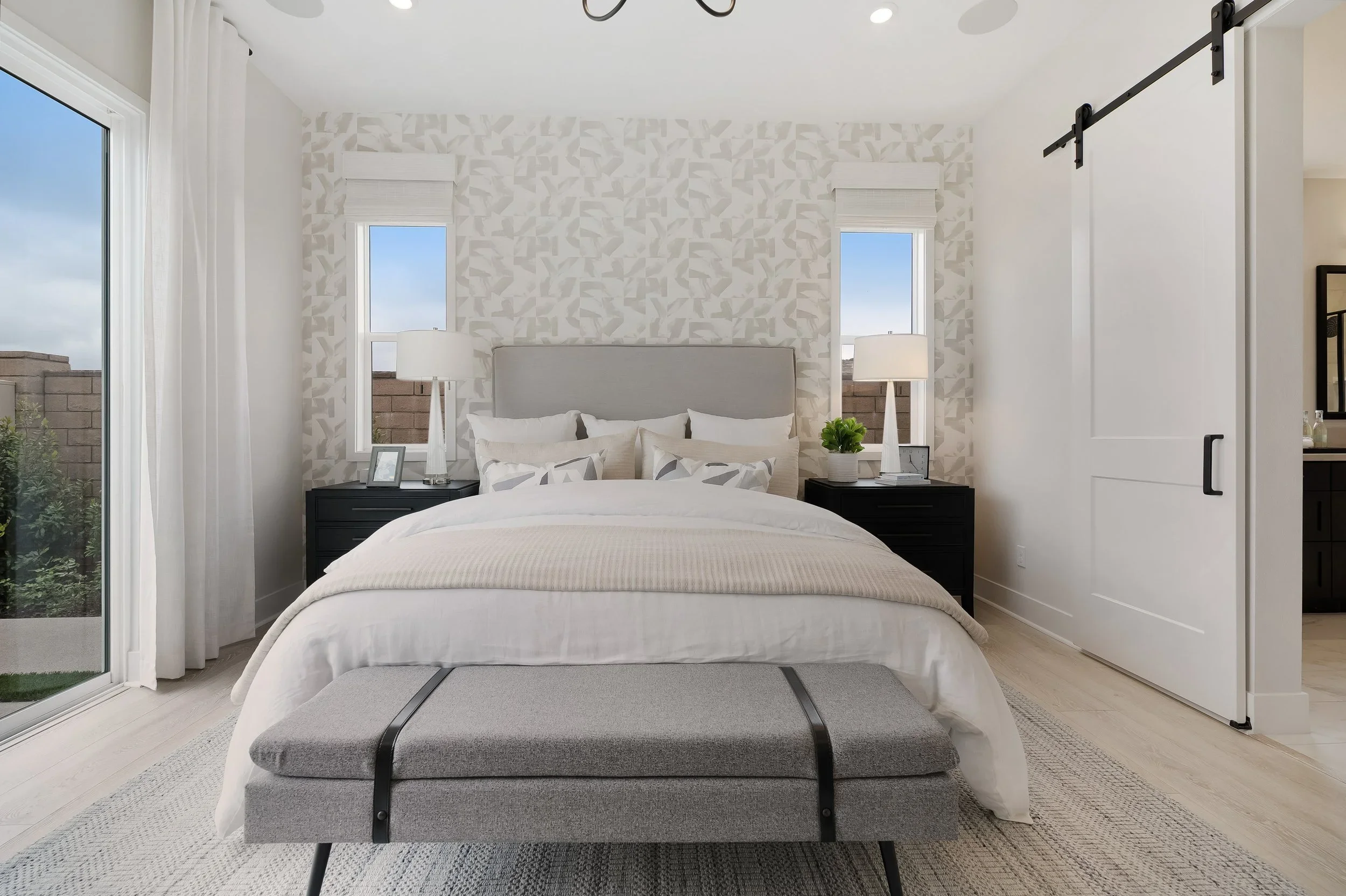 A modern bedroom with a bed, nightstands, lamps, and a bench at the foot of the bed, with windows and a sliding glass door leading outside.