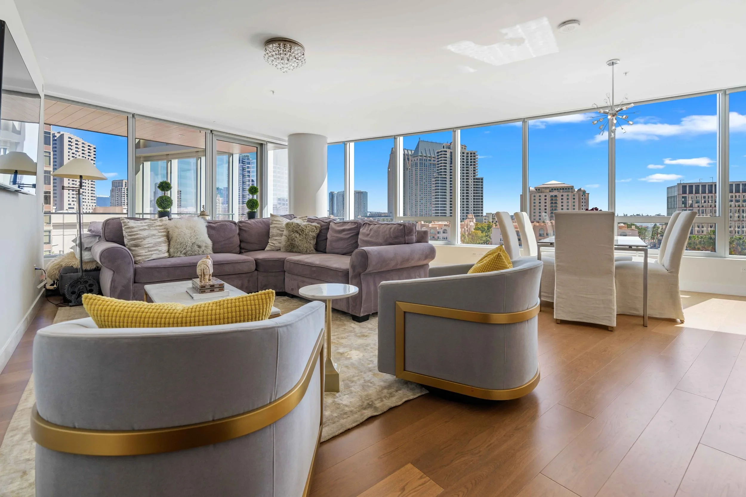 Modern living room with large windows overlooking city skyscrapers, featuring a purple sectional sofa, white armchairs with yellow pillows, and a dining area with six high-backed chairs.