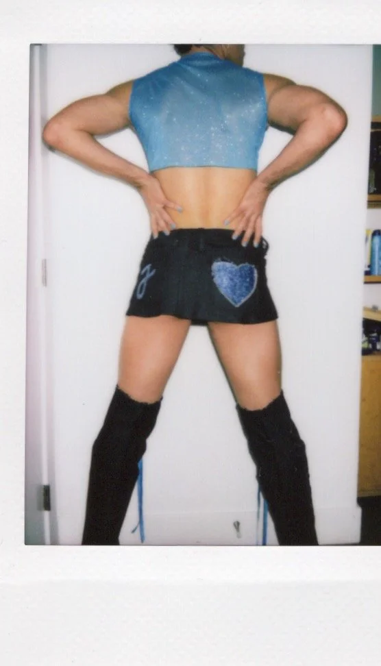 Person wearing a blue crop top, black mini skirt with a heart patch, and knee-high boots, posing with hands on hips.