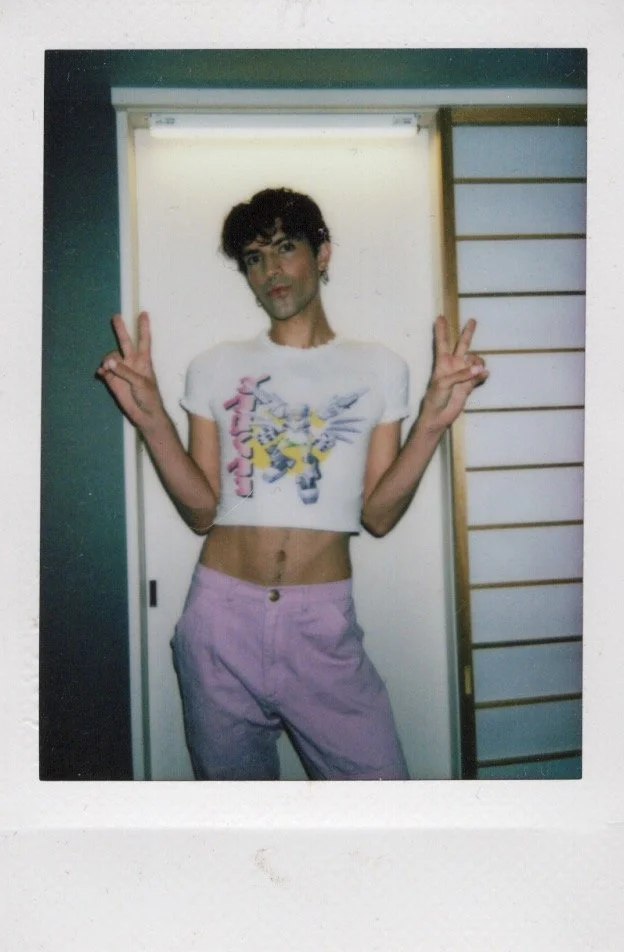 Person posing indoors with peace signs, wearing a cropped graphic t-shirt and purple pants.
